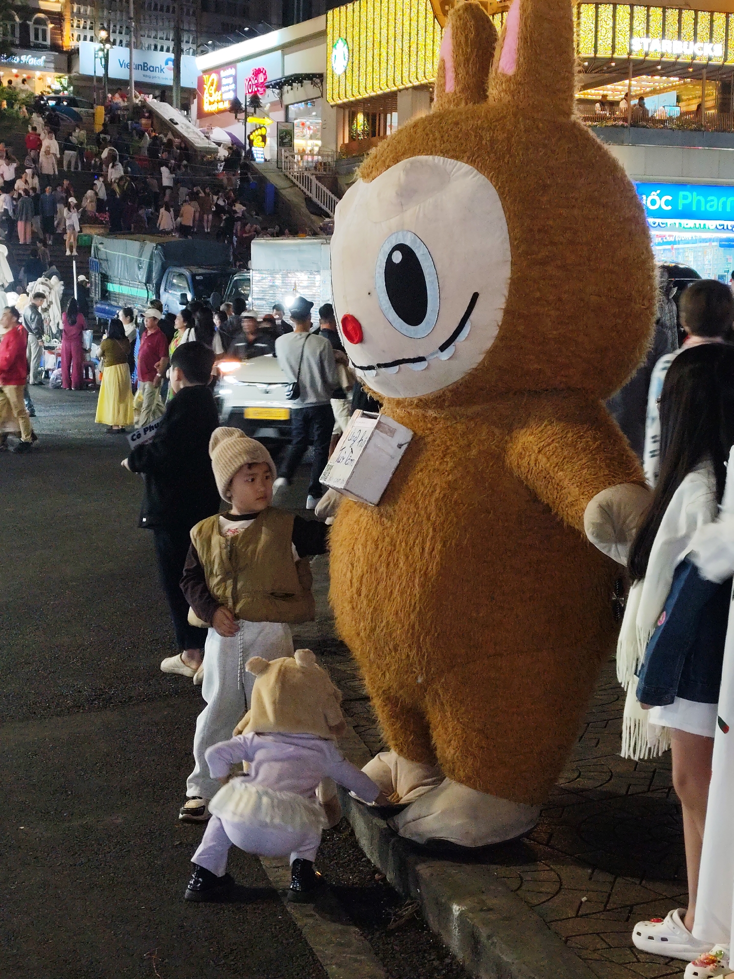 The image depicts a street scene in Da Lat, Vietnam, at night. A large, brown, cartoonish mascot resembling a rabbit stands on a sidewalk next to a curb. Beside the mascot, a young child in a beige knit hat and a brown vest stands near another child, wearing a light purple outfit with a small skirt and a rabbit-eared hood.
In the background, a crowd of people is visible on steps leading up to buildings illuminated by neon signs, including a "VietinBank" sign and a "Starbucks" sign. A vehicle is parked on the street, and a few other people are walking around in the background. The overall environment suggests a lively and crowded public space during the evening.