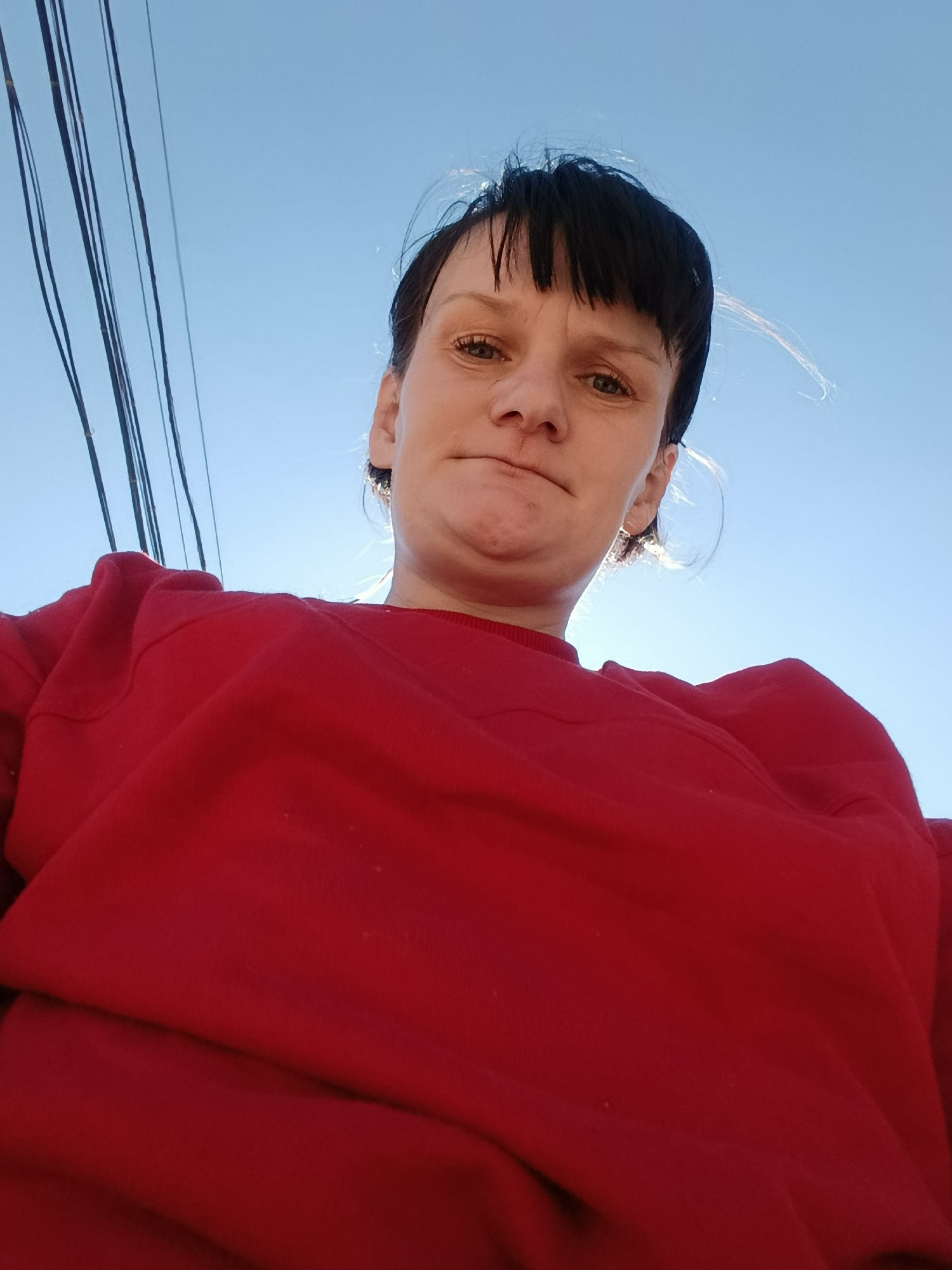A woman with short, dark hair and bangs is seen from a low angle, wearing a red sweatshirt. Above her is a clear, blue sky with a few power lines visible in the upper left corner. The location is the city of Фоки, Russia.