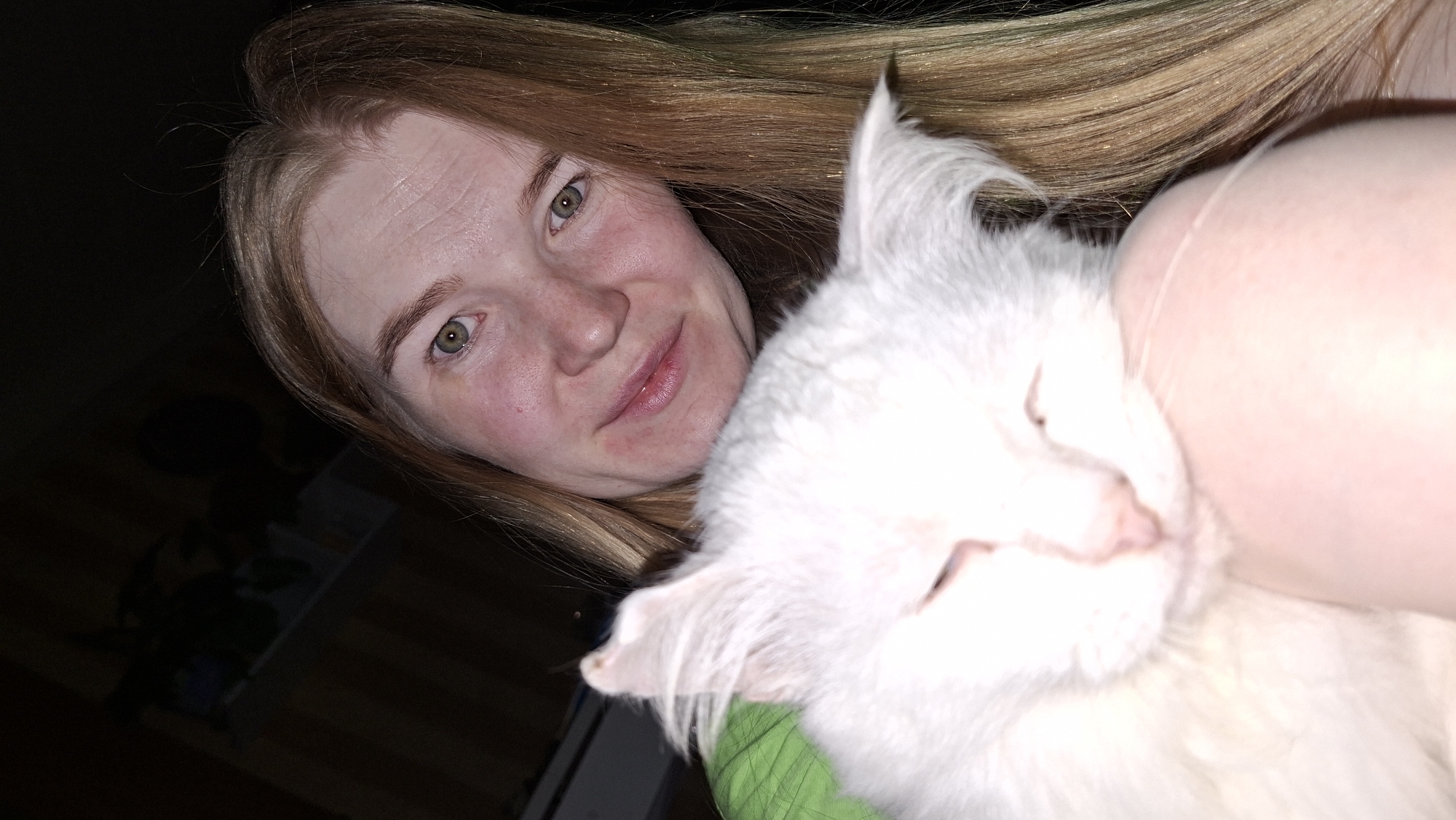 An angled, close-up shot captures a woman and a fluffy white cat in a dimly lit indoor setting, likely in the evening or at night in Большой Букор, Russia. The woman, with fair skin, long light brown or blonde hair, and distinct green eyes, smiles softly at the viewer. Her hair falls around her face, and subtle lines are visible on her forehead.
She is shown in an affectionate embrace with a white cat, whose face is gently snuggled against her arm or shoulder. The cat appears to be sleeping or resting comfortably, with its eyes closed and a hint of its pink nose visible. A portion of green fabric, possibly from the woman's clothing, is visible on the left side of the frame. The background is mostly dark, with some faint reflections on a dark surface visible in the lower left, suggesting the presence of furniture. The image conveys a sense of warmth, peace, and companionship between the woman and her pet. There is no visible text.