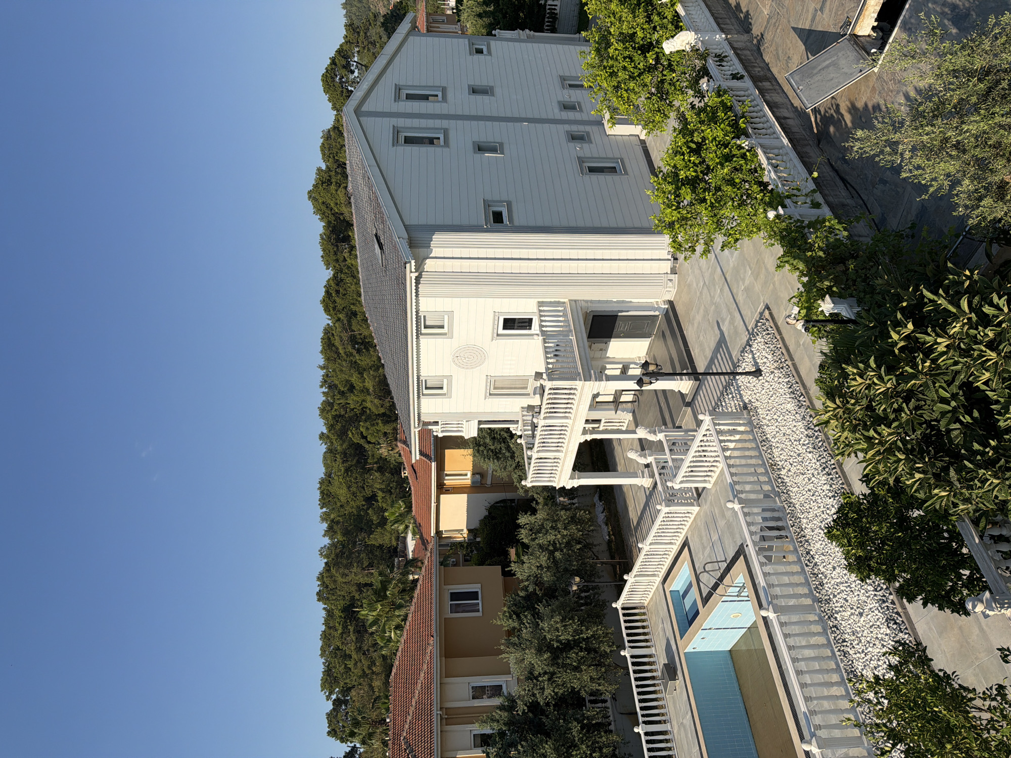 This is an aerial, eye-level, outdoor, daytime, full-shot photograph taken in Unknown, Unknown. The scene depicts a modern villa with a swimming pool in the foreground. The villa is white with a dark grey roof and features multiple stories, balconies, and numerous rectangular windows. A decorative white balustrade surrounds the property, including the pool area. The pool itself is blue with tiled accents and has a ladder. The area around the pool and house is paved with grey tiles and white pebbles. Lush green trees and shrubs are scattered throughout the scene, and a dense forest can be seen in the background. The sky is clear and blue. There are no people visible in the image.