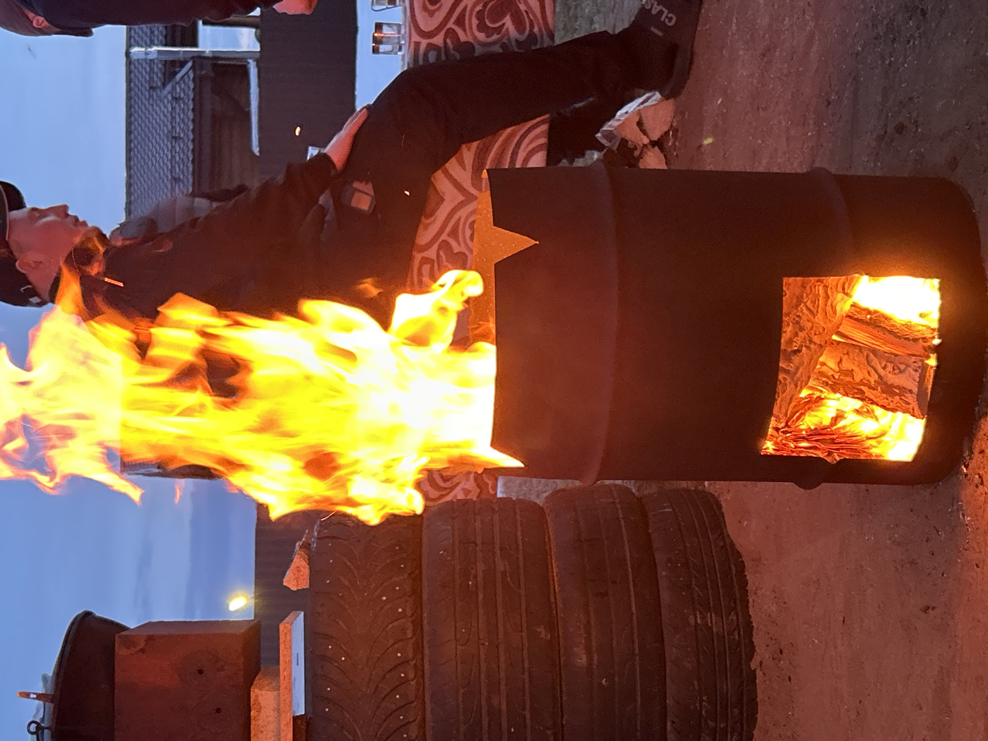 The scene depicts an outdoor evening gathering in Noviy, Russia. Central to the foreground is a modified dark metal barrel serving as a fire pit, from which vigorous yellow and orange flames erupt from the top and a lower rectangular opening where burning wood is visible. To the left, a stack of at least four dark vehicle tires is positioned. Further left, a dark pot rests atop a reddish-brown block. On the right, a male individual wearing a dark baseball cap, dark jacket, dark pants, and dark slide sandals with "CLASS" lettering is seated, his right hand resting on his knee. Part of a second person in a dark cap is visible on the far right edge. Behind the seated individual, a surface covered with a patterned red and white cloth displays several clear glasses. The ground consists of dirt or sand. In the background, a building with a dark tiled roof and a chimney is discernible against a twilight blue sky. A small distant light source is visible in the upper left quadrant.