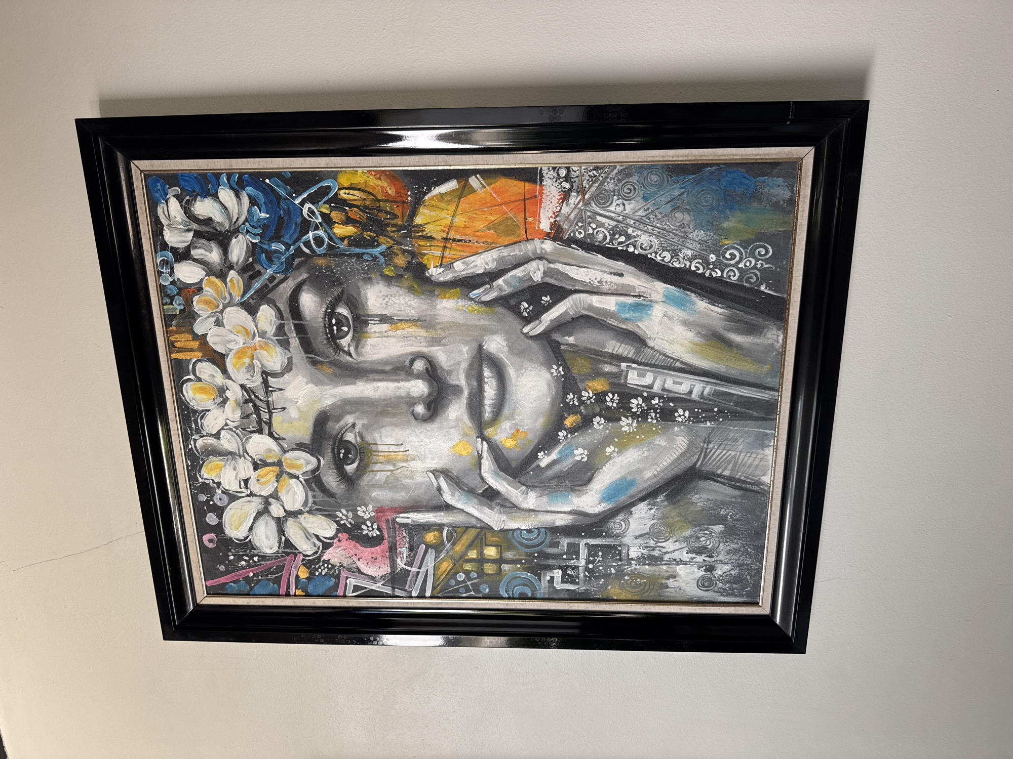 The image shows a painting in a black frame hanging on a white wall. The painting depicts a stylized portrait of a person with flowers in their hair, painted in shades of gray, white, black, and touches of color. The scene is located in Kuta Selatan, Indonesia.
