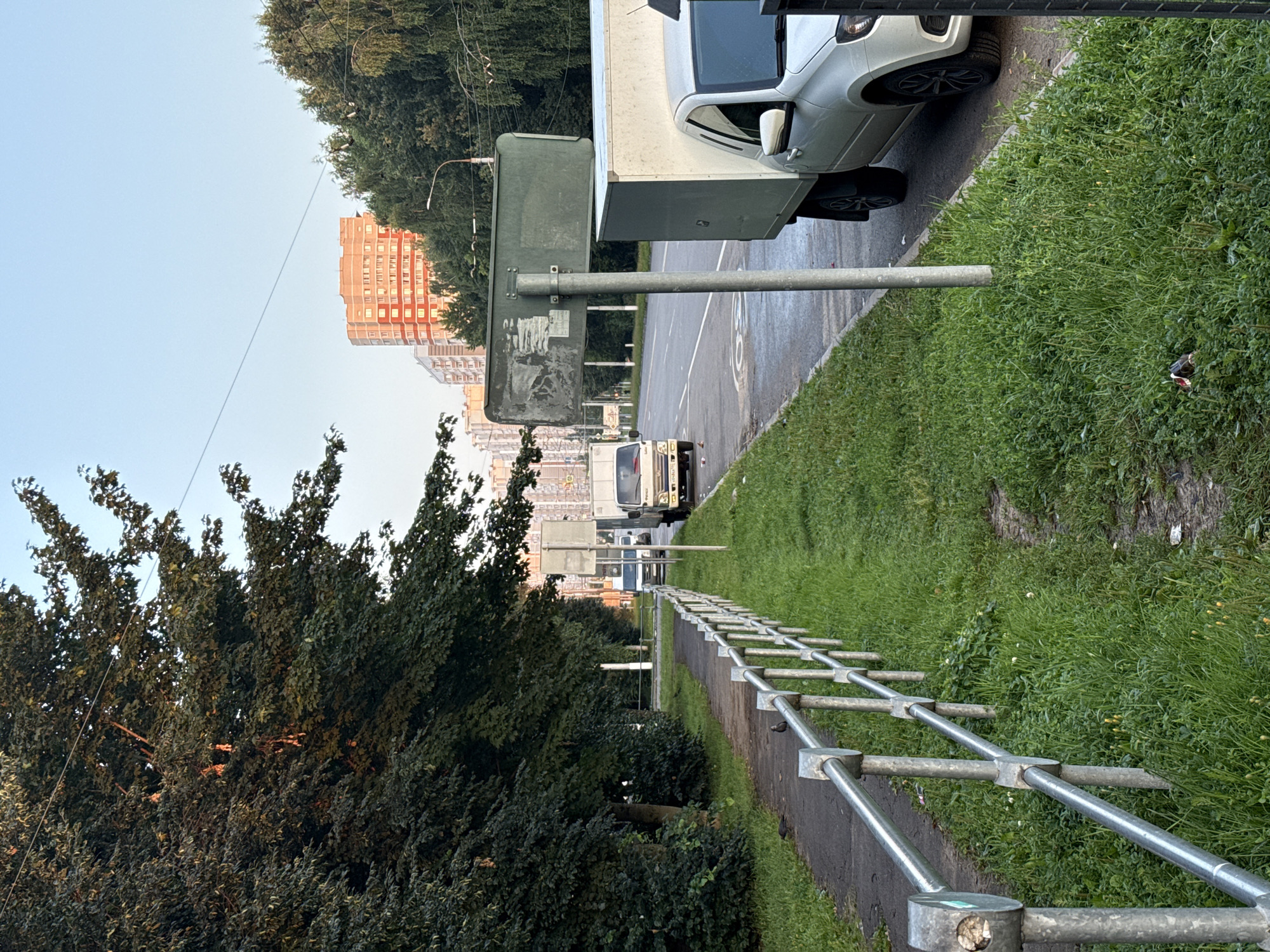 The image captures a street scene in Podolsk, Russia, likely during a late afternoon or early evening due to the lighting. A paved sidewalk runs along the left side of the image, bordered by a silver metal railing. Several small, dark birds are visible on the sidewalk. Beyond the railing, a strip of green grass separates the sidewalk from the road.
The road itself is paved and appears to be a multi-lane street. Two white delivery trucks are parked on the right side of the road. In the foreground on the right, a silver sedan with its rear facing the viewer is parked. A large, dark green sign on a pole is positioned between the sidewalk and the road, partially obscuring a view of the road behind it. The sign appears weathered and has some peeling stickers.
In the background, trees line the right side of the street, and beyond them, tall, multi-story residential buildings are visible. One building in the mid-background is a reddish-orange color and illuminated by the setting sun. The sky is a clear, pale blue, suggesting fair weather. The overall impression is of a typical urban street with some signs of everyday activity.