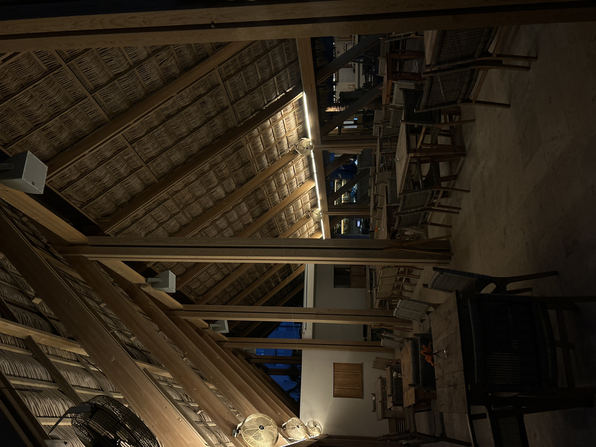 This image depicts the interior of a spacious, open-air or semi-open-air restaurant or bar, likely a palapa structure, at night. The dominant architectural features are a high, pitched thatched roof made of natural fibers and a sturdy framework of large wooden beams. These beams run both diagonally and horizontally, creating a rustic and tropical aesthetic.
Artificial lighting plays a key role in illuminating the space. A strip of warm light runs along one of the prominent wooden beams. Several smaller, round fans with integrated lights are mounted to the ceiling structure, while a larger, caged industrial-style fan is visible in the foreground, also attached to the wooden beams. Rectangular grey speakers, one bearing the "JBL" logo, are mounted on other beams.
The dining area below consists of numerous wooden tables and chairs. The chairs feature woven seats and backs, complementing the natural materials of the roof. The tables are mostly rectangular. The floor appears to be a light-colored, possibly concrete or tiled, surface. No people are present, giving the scene a quiet, serene atmosphere, perhaps indicating after-hours or a lull in activity.
In the background, through openings in the structure, parts of an adjacent building or complex are visible, including white walls and windows with closed wooden shutters. A dark night sky and distant blue lights suggest other outdoor areas or pools, reinforcing the impression of a resort or coastal establishment. The overall ambiance is characteristic of a tropical dining venue in Mazatlán, Mexico, during the evening.