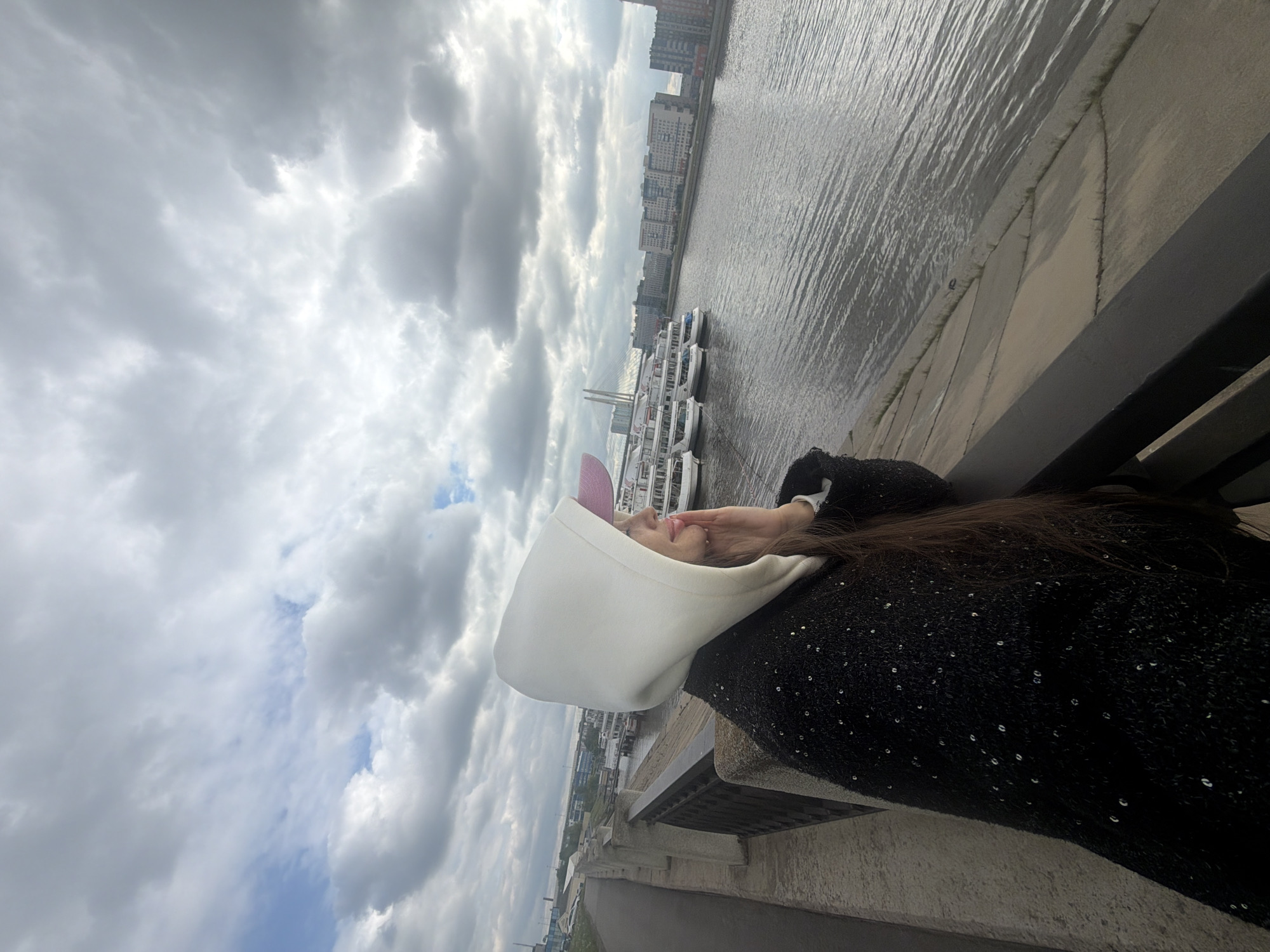 A person is standing on a bridge with a river in the background. The person is wearing a black jacket and a white hoodie with pink cat ears. There are several boats docked along the riverbank. The sky is cloudy. The Vantovy Bridge is visible in the background, and it is likely that the location is Saint Petersburg, Russia.