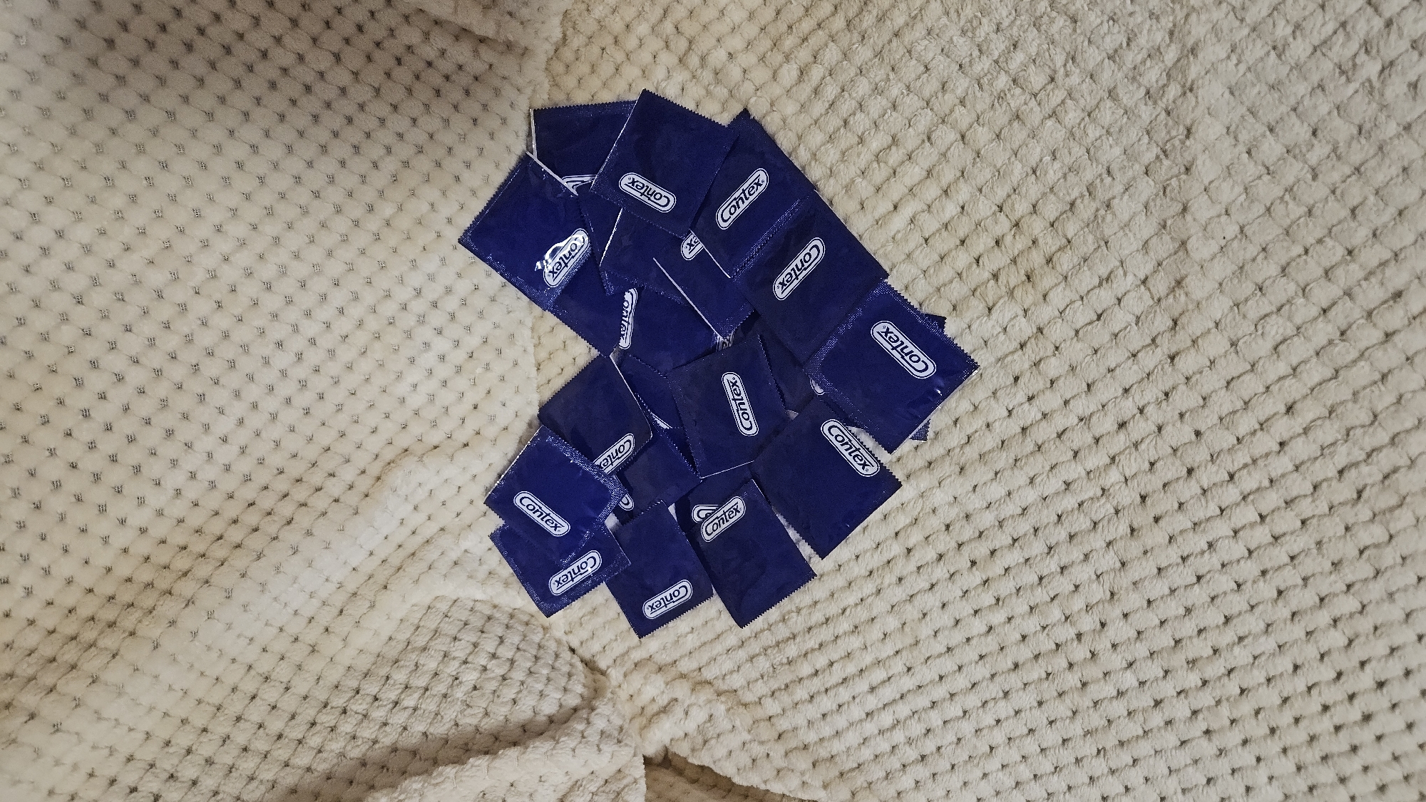 The image shows a collection of approximately 15 blue condom wrappers, each labeled "Contex", arranged in a roughly square cluster on a textured beige blanket or throw. The blanket has a dense, repeating pattern of small squares or waffles. The location is not discernible from the image.