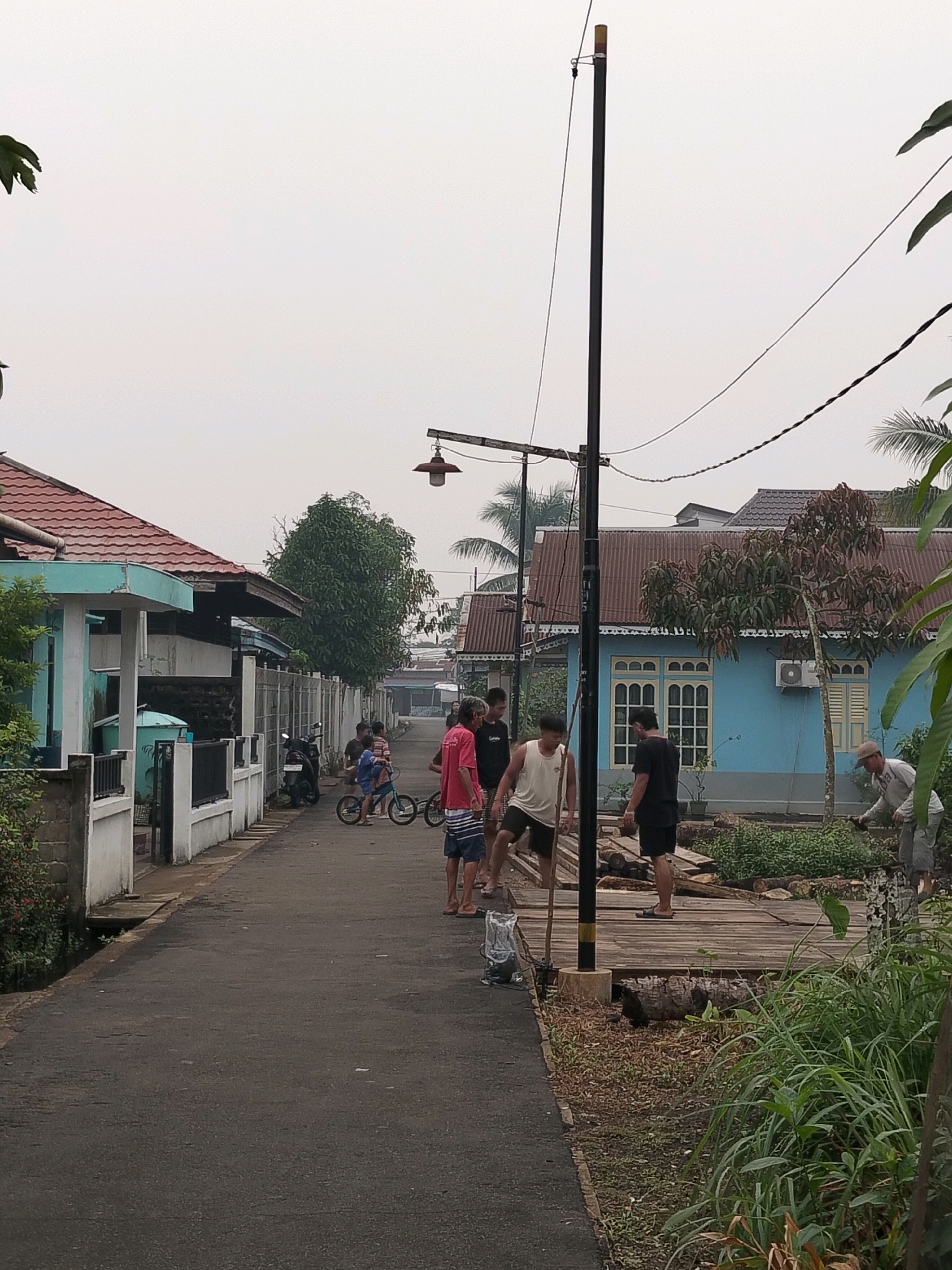 The image depicts a street scene in Arang Limbung, Indonesia, under an overcast sky. Several individuals, predominantly young males and at least one older male, are present. Some are casually standing, while others are engaged in activities such as riding bicycles. A man in a light-colored shirt and hat appears to be working near some greenery and wooden planks.
The environment includes residential buildings with tiled roofs and distinct architectural features, such as arched windows on a blue house. A paved road runs through the scene, with a small canal or drainage ditch running alongside the houses on the left. Various objects are visible, including motorcycles, bicycles, wooden logs, and electrical poles with wires. Vegetation, including trees and tall grasses, is interspersed throughout the scene. The overall atmosphere suggests a typical residential neighborhood.