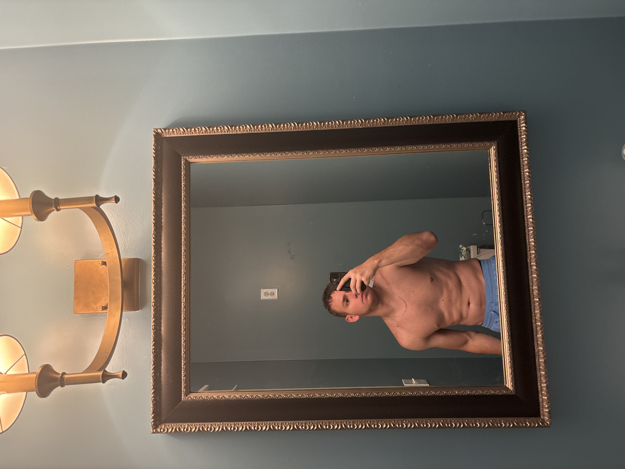 The image shows a person taking a mirror selfie in a bathroom. The person is a shirtless male with muscular build, wearing blue boxer briefs. He is holding a smartphone in his right hand, angled to capture his reflection. The bathroom has teal-colored walls and a large, ornate mirror with a dark frame. To the left of the mirror, there is a golden wall sconce with two lights. The lighting appears to be artificial, suggesting it could be nighttime or an indoor setting with the lights on. The overall mood is casual and self-observational. No visible text is present in the image. The location context of Panama City Beach, USA, is provided, which could imply the setting is a residential bathroom or hotel.