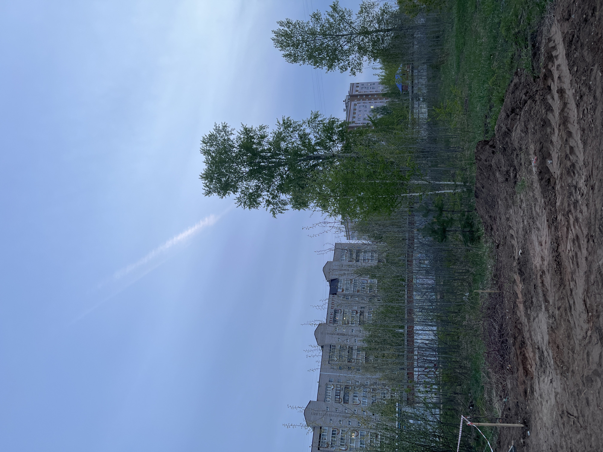 The image shows a dirt mound in the foreground. Behind it is a row of young trees in front of an apartment building. More trees and another apartment building are in the background. The sky is blue with a contrail. The location appears to be in a city, likely Chaikovsky, Russia, given the context. There are no visible people. A security barrier is visible on the right side of the mound.