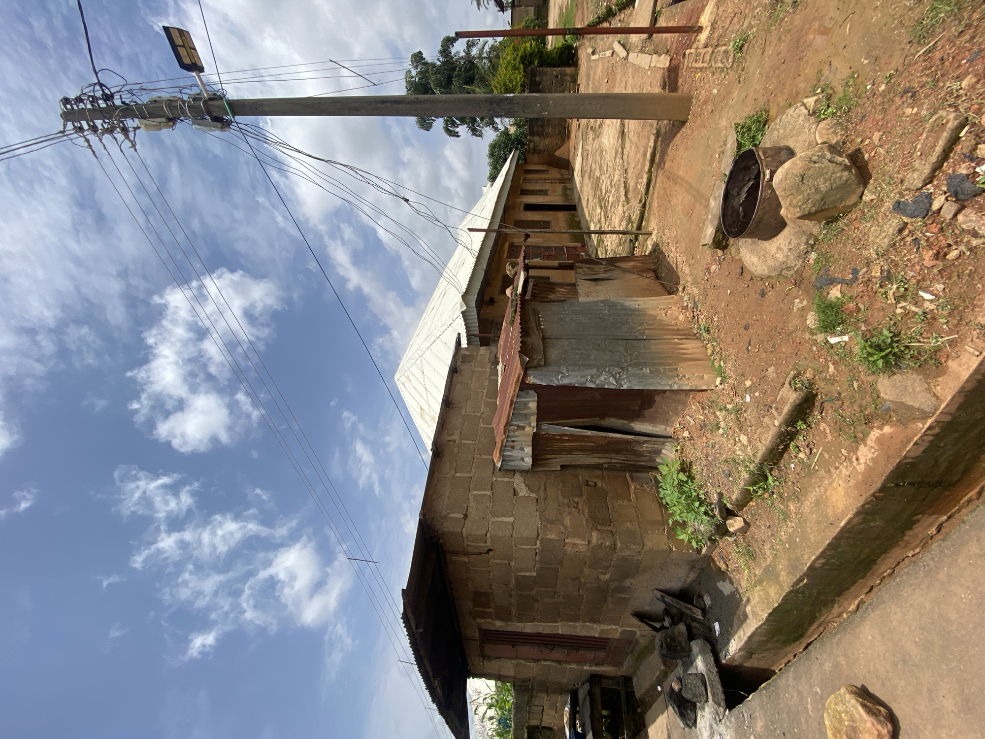 The image depicts an outdoor scene in Gura Top, Nigeria, featuring several structures and a utility pole. In the foreground, a concrete drainage channel runs along the left side of the image. Adjacent to the drainage is a small, roughly constructed building made of cement blocks with a corrugated metal roof. To the right of this building is another smaller structure, also with a corrugated metal facade.
Further back, a larger building with a white, sloped corrugated metal roof is visible, stretching horizontally across the image. This building appears to be an extended structure with multiple doors or windows. A concrete patio or ground covers the area in front of this building.
Dominating the right side of the image is a concrete utility pole adorned with numerous electrical wires and transformers. A street light fixture is attached to the pole. The ground is composed of reddish-brown soil, scattered with rocks, debris, and patches of green vegetation. The sky above is blue with scattered white clouds, indicating daylight. No people are present in the scene.