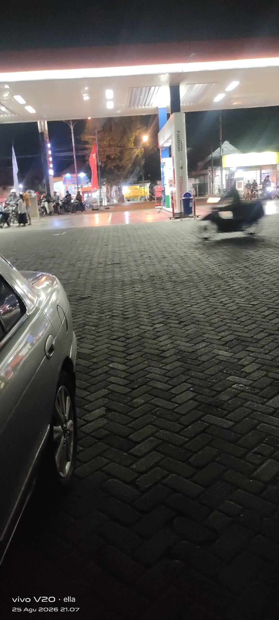 This is a nighttime photograph taken at a gas station in Tanjungsari, Indonesia. The foreground is dominated by the paved ground, a herringbone pattern of dark bricks. To the left, the rear portion of a silver sedan is visible, its wheel and part of its bodywork illuminated by ambient light. In the middle ground, a gas pump with a blue and white structure is prominent, featuring the "Fastron" logo. Several motorcycles and people are gathered around the pumps, with some vehicles in motion, creating motion blur. The background shows the illuminated canopy of the gas station and buildings with various signs and lights, indicating commercial activity. The overall scene depicts a typical bustling gas station environment at night.