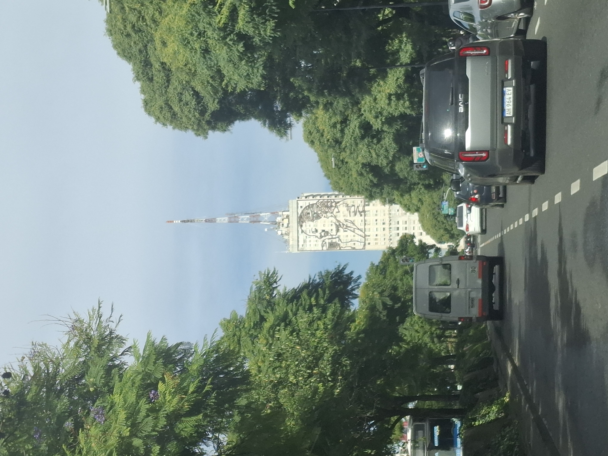 The image shows a street scene in Buenos Aires, Argentina, likely taken from inside a moving vehicle. The focus is on the Avenida 9 de Julio, as indicated by the distinctive building in the background – the Ministerio de Desarrollo Social building with the portrait of Eva Perón. A tall antenna or transmission tower is visible near the building.
The image captures vehicles on the avenue, including a BAIC car, a delivery van, and several other cars in the traffic. The road is marked with lane lines. Trees line the avenue, partially obscuring the view of the buildings and sky. The sky is blue with some visible haze.