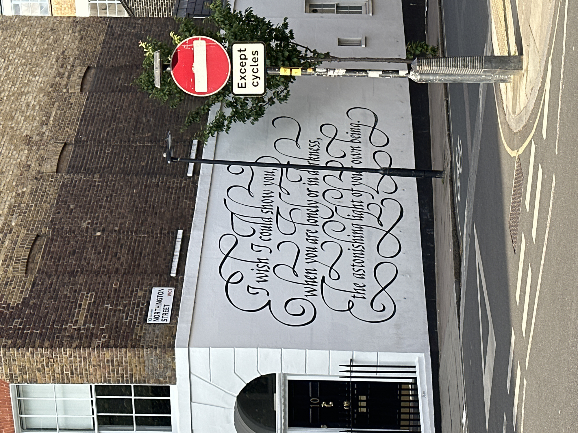 The image shows a street in London, United Kingdom. A white building with a large quote in black cursive script is visible, reading, "I wish I could show you, when you are lonely or in darkness, the astonishing light of your own being." A black front door with the number "10" is beneath the quote. Above the building, a street sign reads "NORTHINGTON STREET WC1" on the building. A "no entry except cycles" sign sits on a pole near the building. The street shows crosswalk markings. The image is rotated clockwise by 90 degrees.