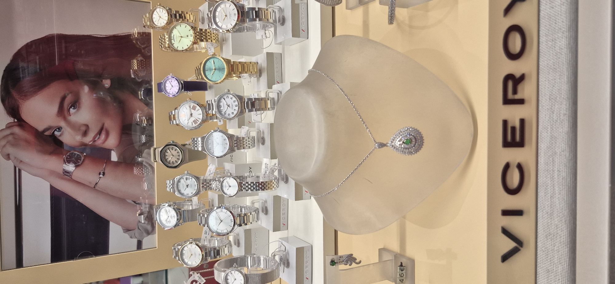 The image shows a display of Viceroy brand watches and jewelry in a store located at Rua da Eça de Queirós, 23, 4500-308 Póvoa de Varzim, Portugal. The display features multiple watches of various styles and colors, arranged on white stands. In the foreground, a mannequin bust showcases a silver necklace with a teardrop-shaped pendant containing a green stone. To the left, a price tag displays "46€" next to a pair of dangling earrings with a green stone. A large advertisement poster of a woman wearing a watch is visible at the top of the display. The overall lighting is bright, highlighting the merchandise.