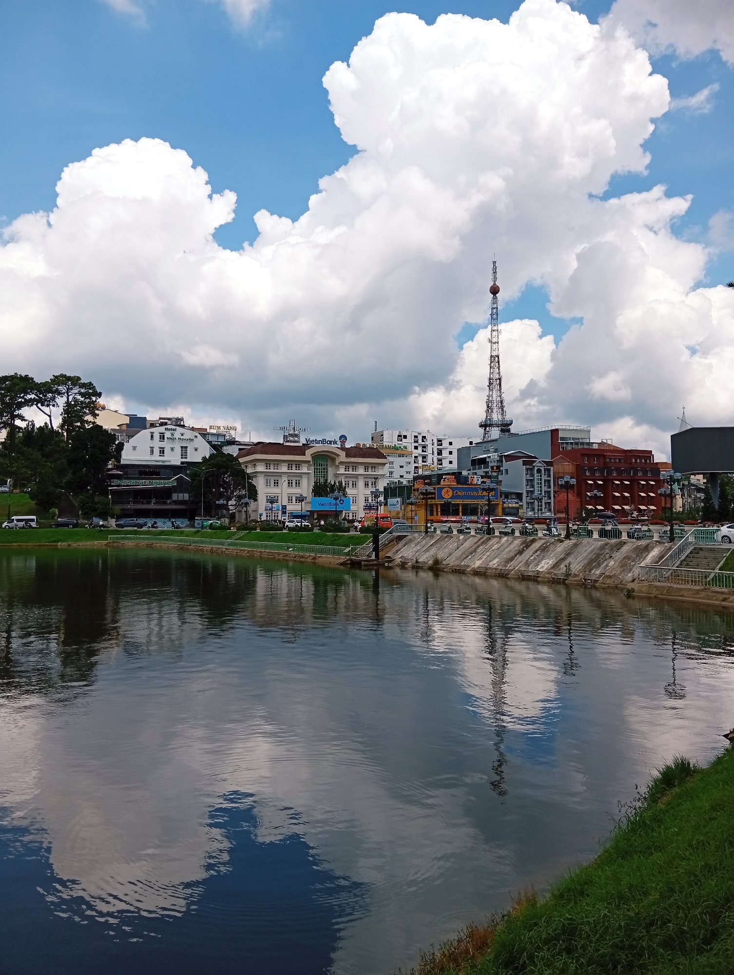 The image captures a wide view of Xuan Huong Lake in Da Lat, Vietnam, on a partly cloudy day. The lake's calm surface reflects the sky and the buildings lining its edge. On the far bank, a variety of buildings are visible, including hotels and commercial establishments, with a prominent telecommunications tower rising above them. Trees, including tall pines, line the embankment on the left. A concrete walkway and steps lead down to the water's edge, and on the right, a grassy slope slopes down towards the lake. Several cars are parked along the road beside the buildings, and a few people are visible walking or standing near the shops. The overall impression is of a scenic urban landscape with natural elements.