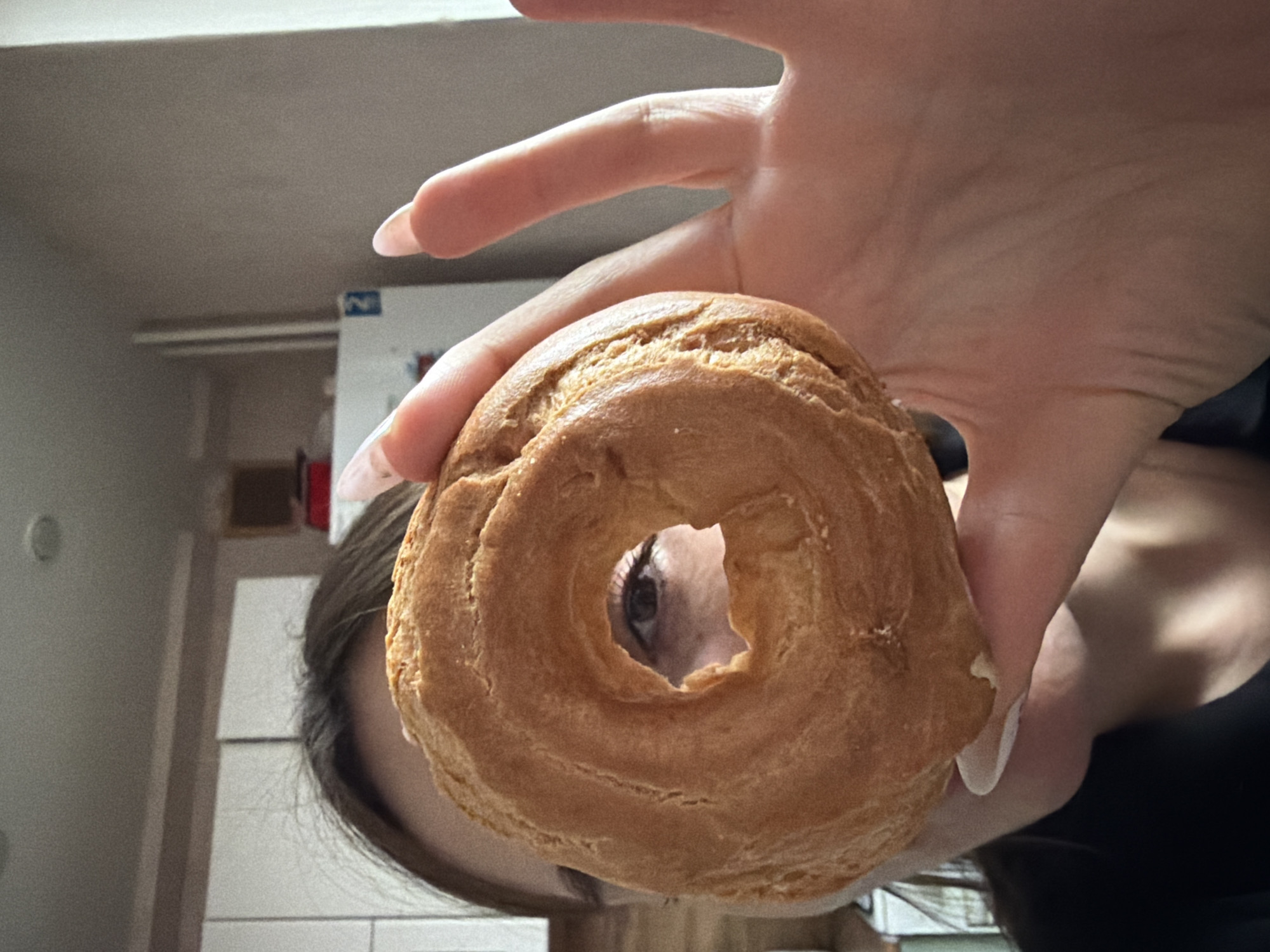 A person with long, manicured fingernails is holding a ring-shaped pastry at eye level. Through the hole in the pastry, the person's eye is visible. The background shows a portion of a room, including a refrigerator, part of a wall, and a glimpse of another room with a white wall. There is no indication of this image being taken in Izhevsk, Russia.