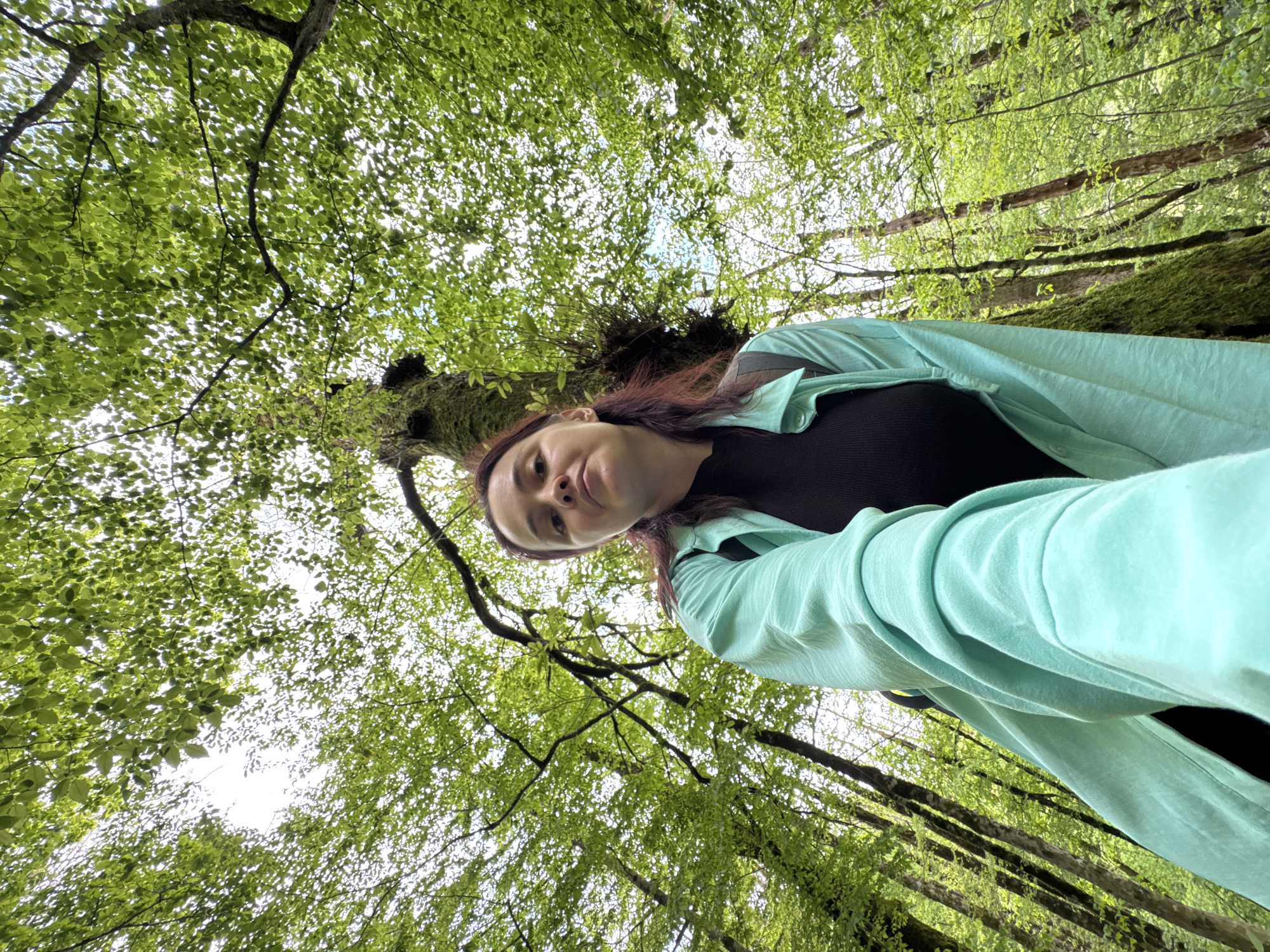 A woman is in a forest in Krasnaya Polyana, Russia. The camera is oriented upward, capturing a low-angle perspective. The woman is wearing a black shirt and a light blue shirt, and has reddish-brown hair. Trees surround the woman, with green foliage visible through the branches, with moss on some of the trunks.
