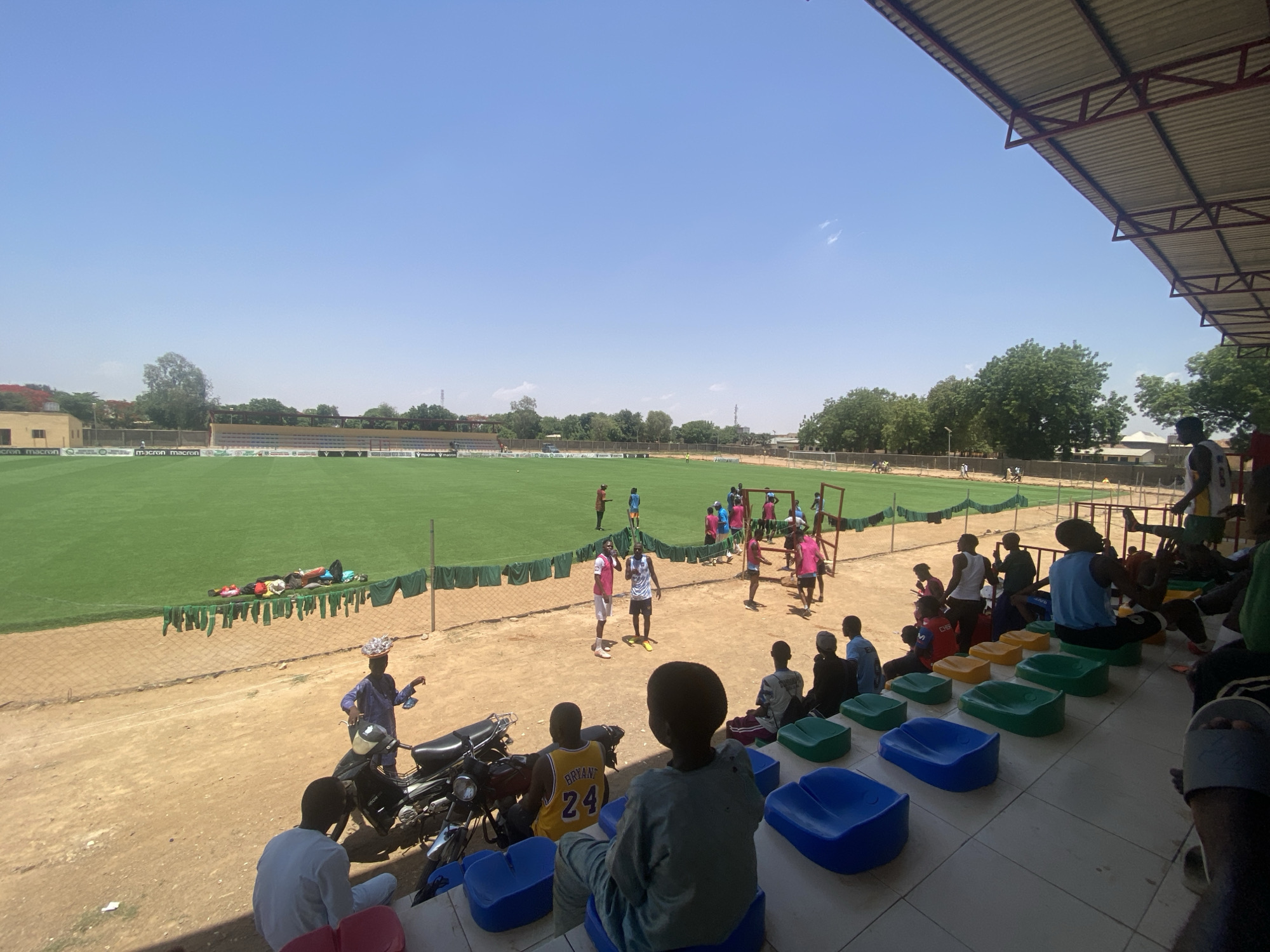 The image shows a soccer field and stadium in Tudun Wada, Nigeria. The sky is clear and blue. In the background, a green soccer field is visible with players practicing. A low fence borders the field. Behind the fence, some clothing is hanging to dry. In the foreground, stadium seating is visible with a mix of colored seats (blue, green, yellow, red). Several people are seated in the stadium, observing the activity on the field. Some individuals are wearing jerseys, including a yellow jersey with the number 24. A black motorcycle is parked near the seats. A person is standing near the motorcycle, wearing a traditional headdress.