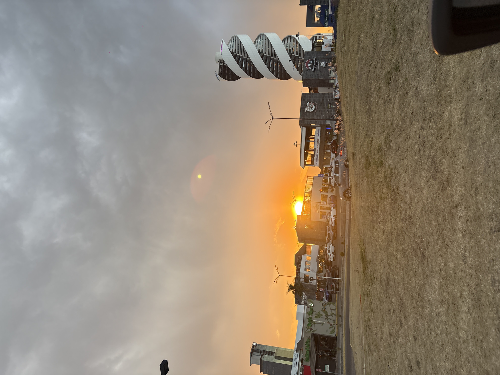 This image depicts an urban landscape at sunset in San Bernardino Tlaxcalancingo, Mexico, under a partially cloudy sky. The sun, a bright orange orb, is low on the horizon, positioned centrally to the right, and casts a prominent golden hue across the lower sky. Above the horizon, the sky transitions to overcast grey clouds.
The scene features several commercial structures and a distinctive modern high-rise building. On the left, a multi-story building with "Italian Coffee" signage is visible. To its right, a lower building displays "Pasteleria". Further right, the sun obscures some details, but a complex of businesses including "Los Originales Tacos de Pescado y Camaron" and "Rocky Mountain" is identifiable. The most prominent structure on the right is a tall, white, multi-story building with a distinctive helical or spiral architectural wrapping. Another modern, taller building is partially visible on the far left.
A paved road or street runs horizontally across the midground, with numerous vehicles parked along its edge and within lots, including several white sedans, a dark SUV, and a silver sedan in the lower left. Multiple streetlights are active or present. The immediate foreground consists of a large expanse of dry, brown grass. A portion of what appears to be a dark car interior or dashboard is visible in the lower right corner.
