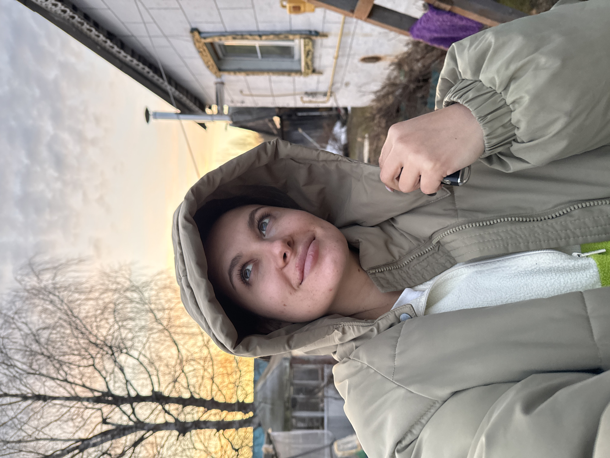A medium shot depicts a woman in a hooded jacket, holding a key in her right hand. She is outside, with a house and bare trees in the background. The sky is partly cloudy with hints of sunset.