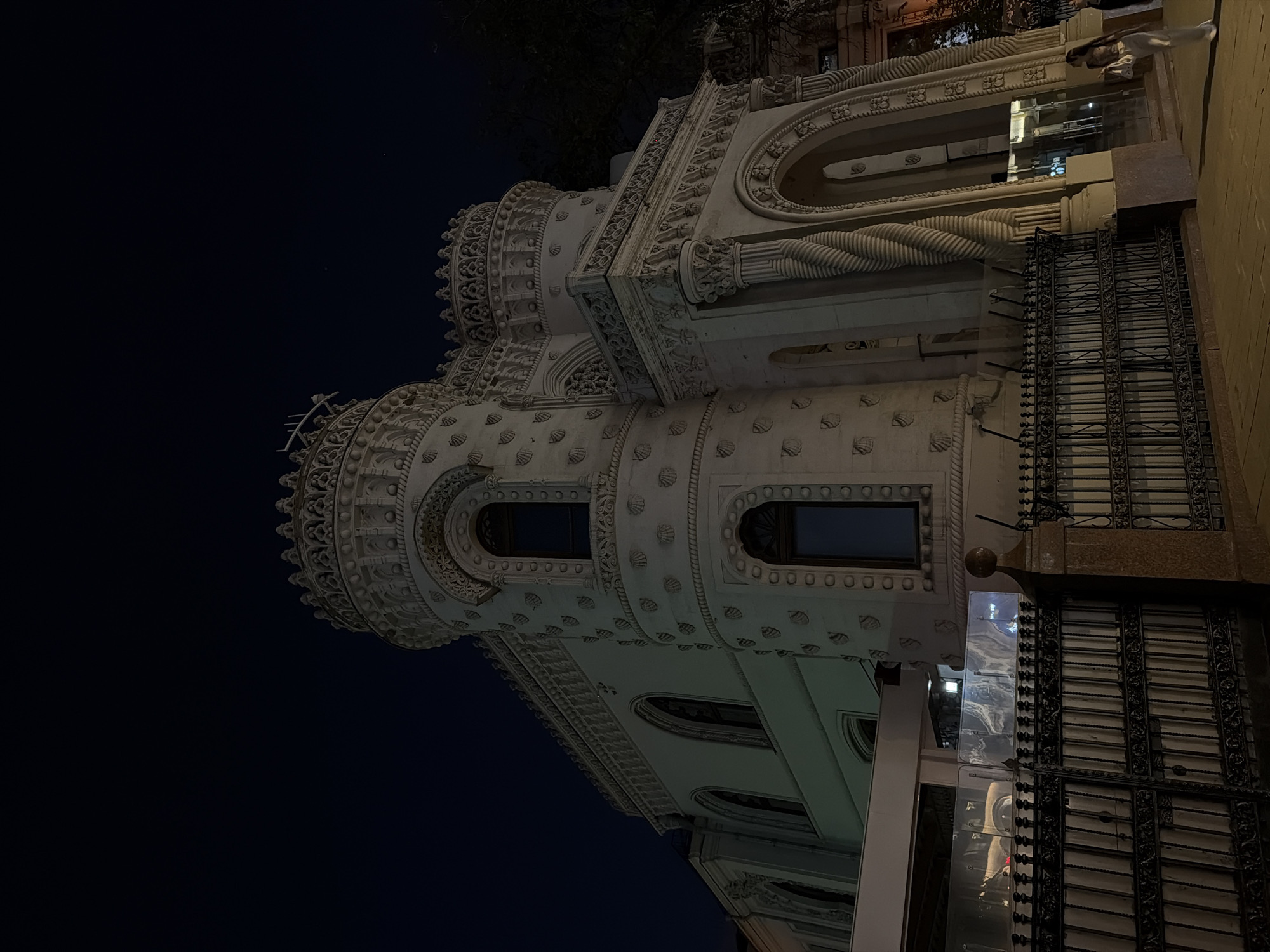 The image depicts the exterior of the Igumnov House, a building in Moscow, Russia, at night. The building is predominantly white and features ornate detailing, including shell-shaped patterns and decorative moldings. The architectural style is distinctive, with a tower-like structure and arched windows. A wrought-iron fence runs along the base of the building. The sky is dark, indicative of nighttime.