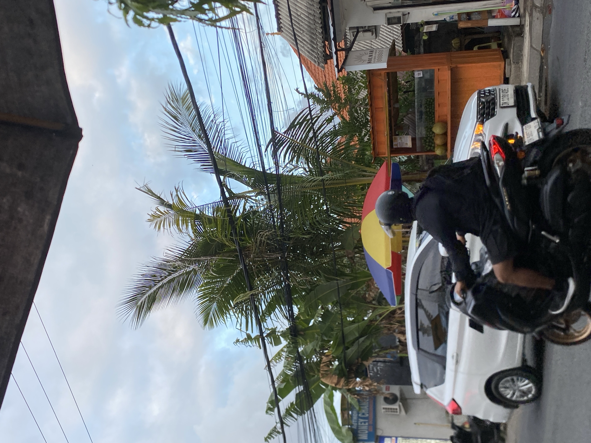 The image depicts a street scene in Mengwi, Indonesia, with a focus on a person riding a motorcycle in the foreground, traveling on a paved road. The rider is wearing a black helmet and black clothing. Behind the motorcycle, a white car is stopped, possibly in traffic. To the right of the car is a wooden stall displaying fruits, with an umbrella over it. Further back, there are palm trees and banana trees, suggesting a tropical environment. Buildings with red-tiled roofs are visible in the background, along with numerous electrical wires overhead. A sign that says "MONEY CHANGER" is visible on a building to the left. The sky is partly cloudy.