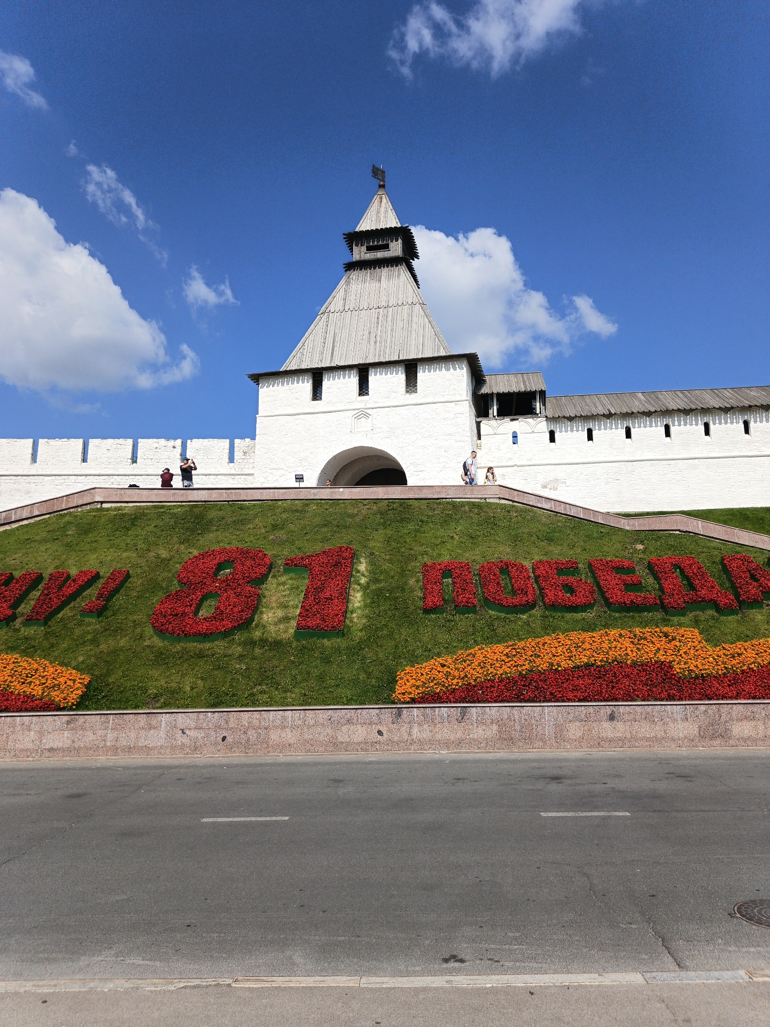 The image shows a sunny day in Kazan, Russia, with a clear blue sky dotted with fluffy white clouds. In the foreground, a road with white lane markings is visible. Behind the road, a vibrant hillside is decorated with a large floral arrangement forming the number "81" in large red flowers, followed by "ПОБЕДЫ" (meaning "Victory") in red flowers. This floral display is situated in front of a white stone wall with arched entrances and wooden roofs, characteristic of Kazan's historical architecture, possibly part of the Kazan Kremlin complex. Several people are visible on the elevated walkway above the floral display, with some taking photos and others walking. The overall scene depicts a celebratory outdoor event or commemoration in a city known for its rich history and cultural landmarks.