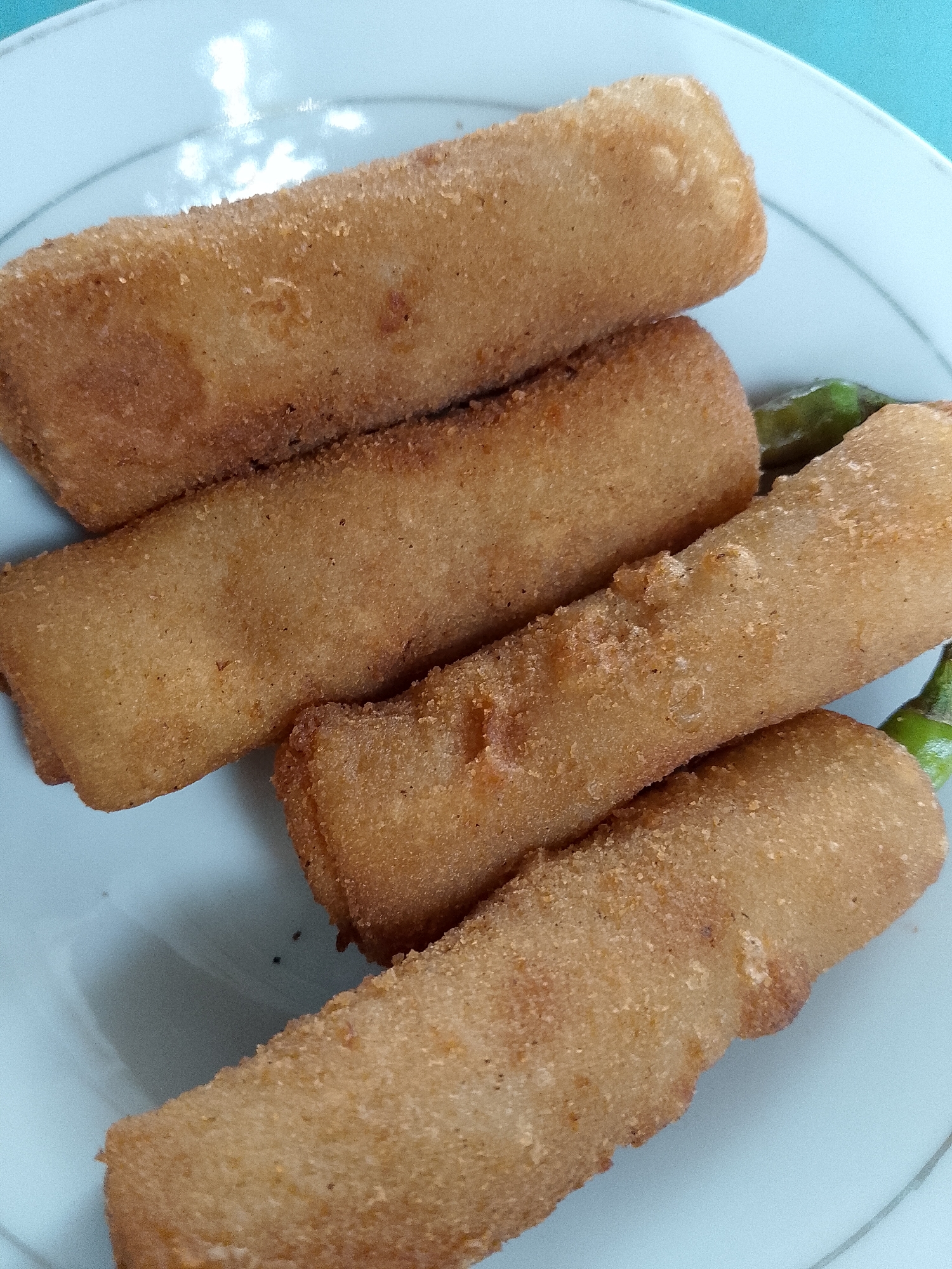 A close-up, overhead shot shows four golden-brown, cylindrical fried snacks arranged diagonally on a white plate. Some green chili peppers are visible peeking out from behind the snacks on the right side of the plate. The snacks have a slightly textured, breaded exterior. The background is a plain light blue surface. The image appears to have been taken indoors, possibly in a home or a small eatery in Lhokseumawe, Indonesia, given the commonality of such fried snacks and chili peppers in Indonesian cuisine.