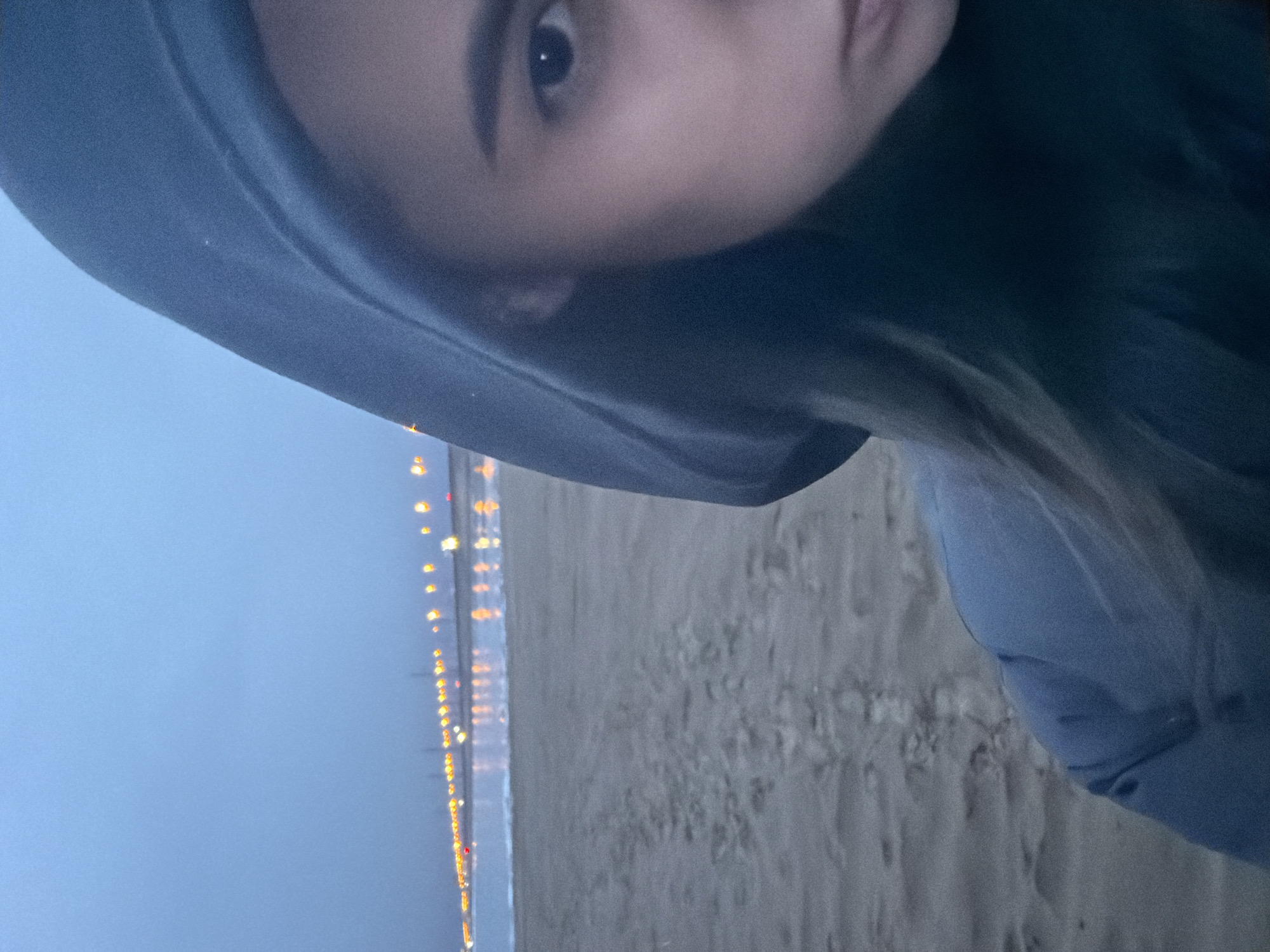 This vertical image, taken at dusk or night, features a person on a beach in Noviy, Russia. The upper right portion of the frame shows a close-up of a person, likely female, looking towards the camera. Her right eye, eyebrow, and part of her face are visible, partially framed by the dark hood of a jacket or hoodie she is wearing. Her expression appears neutral or slightly reflective.
The background depicts a sandy beach or shore beside a body of water, possibly a sea or large lake. A prominent line of warm-toned lights, likely from a distant city, pier, or coastal establishments, stretches across the horizon, creating a luminous boundary between the water and the dark sky. The sky itself is a deep, muted blue, indicating the low light conditions of evening or night. The sand on the beach exhibits texture and appears damp in places. The overall atmosphere is quiet and serene, suggesting a moment of solitude by the water in cool weather. No text is visible in the media.