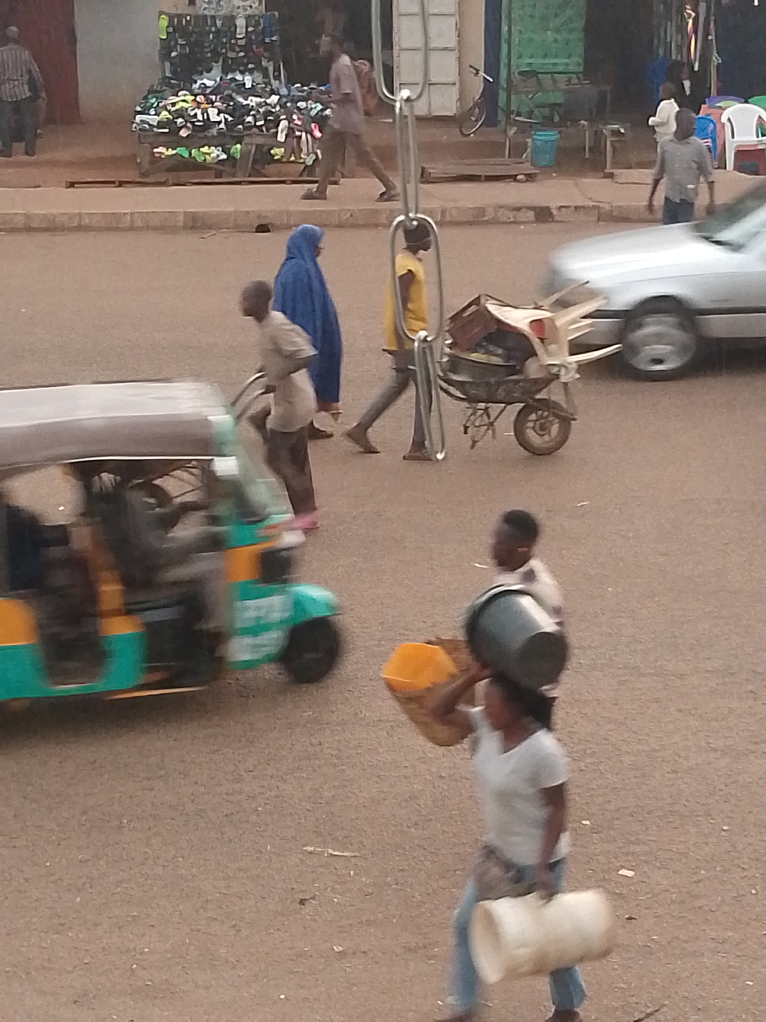 In Bukuru, Nigeria, a busy street scene unfolds with an auto-rickshaw in motion. To the left, a person is seated within the auto-rickshaw. A woman in a blue headscarf walks past. A man in a light-colored shirt is near the auto-rickshaw. In the foreground, a woman carries a large black bucket on her head and a large white container in her arms, while another woman carries a yellow object on her head. In the background, a stall displays numerous shoes and sandals. A silver car is visible on the right side of the image, and a person pushes a wheelbarrow filled with items. The ground is paved with asphalt.