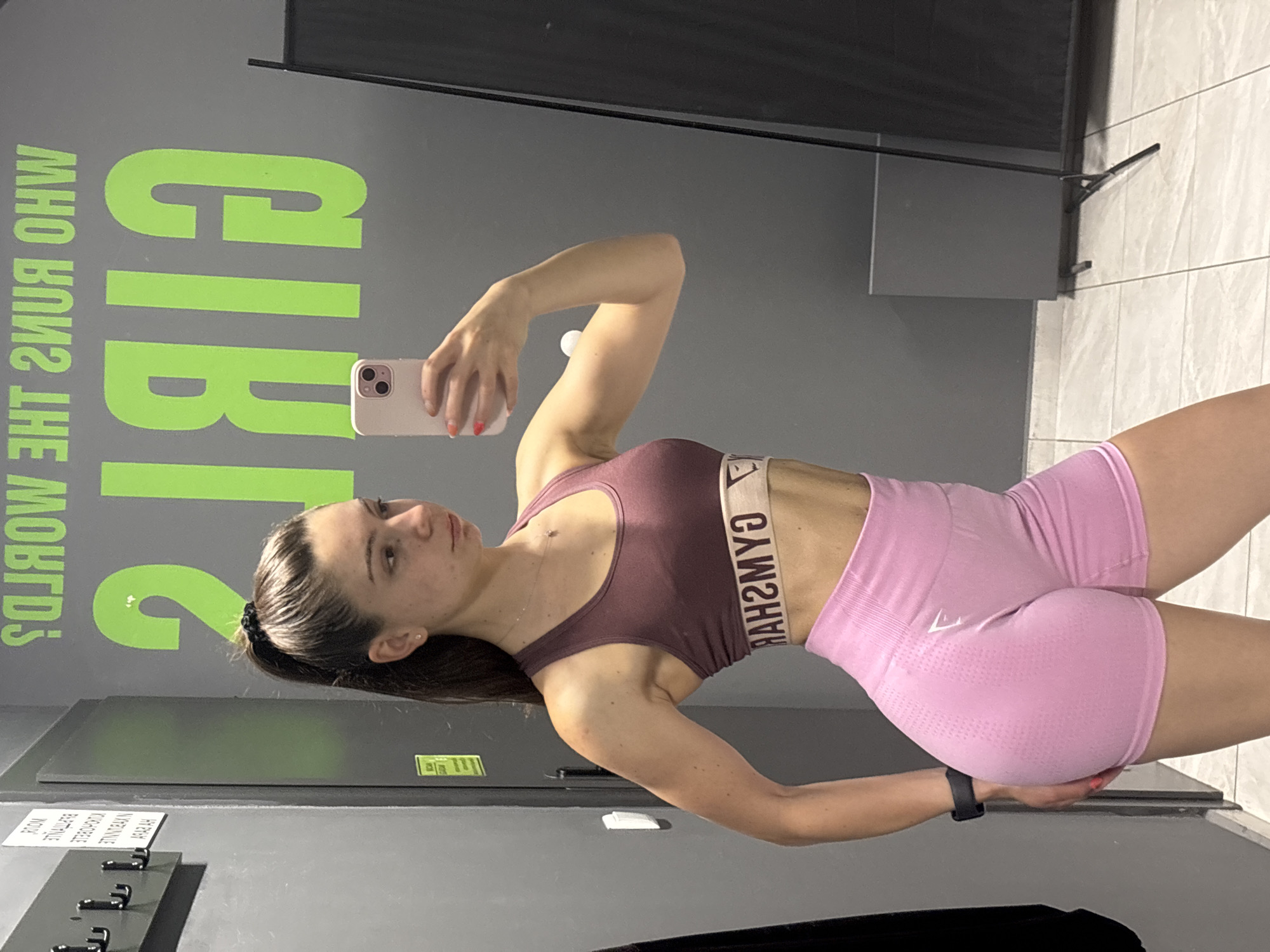 A woman is taking a selfie in what appears to be a gym changing room or a gym area. She is wearing a maroon sports bra and pink shorts, both featuring the brand name "GYMSHARK". She is holding a phone up, angled to capture her image in the mirror. The background includes a gray wall with a large, green text graphic that reads "GIRLS WHO LIFT". A metal clothes rack is visible on the wall near a locker. The floor is tiled and a black object is on the floor. The scene does not suggest a specific location in Varna, Bulgaria.
