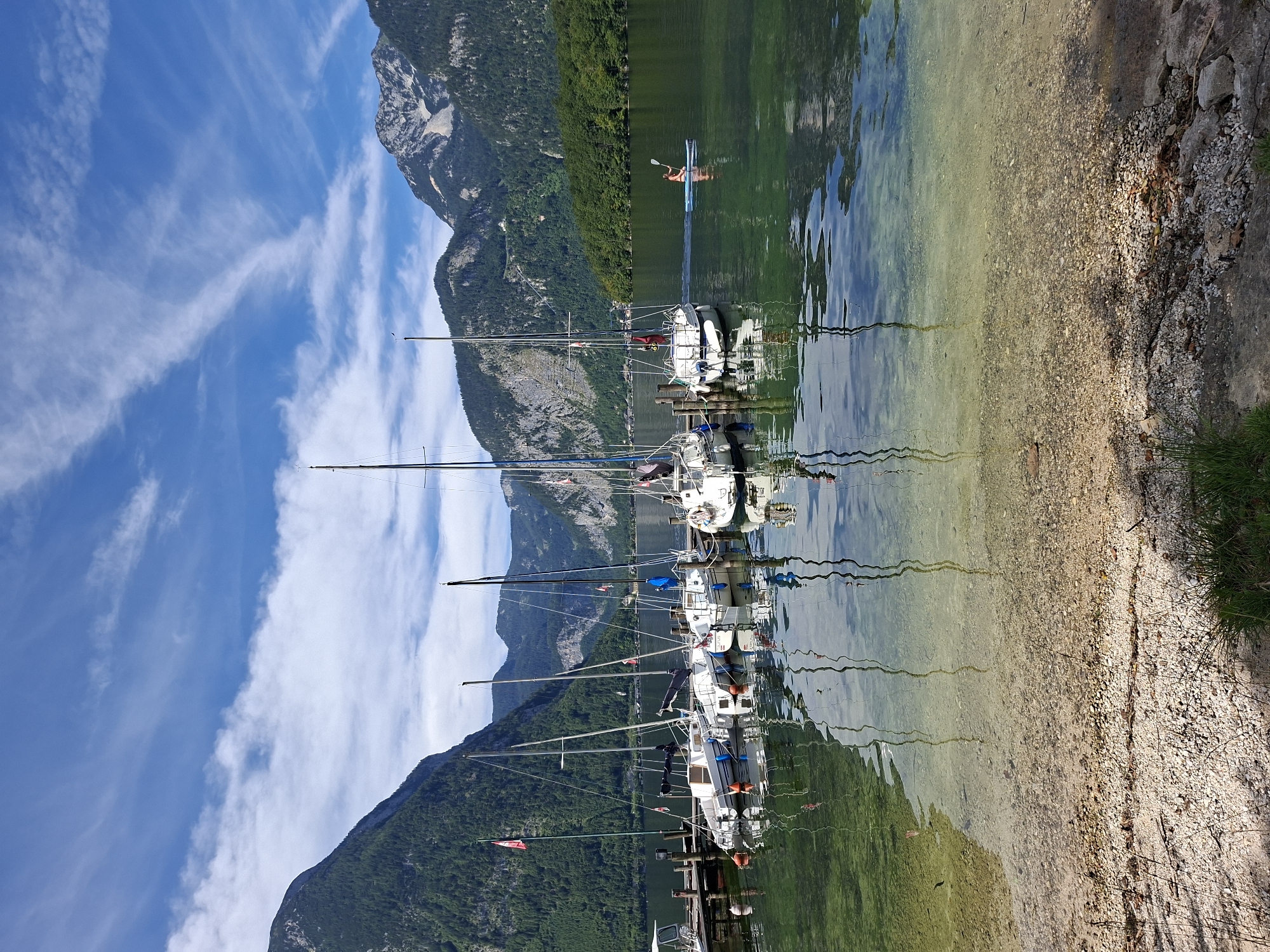 A wide shot of a lake with sailboats docked at a pier on the left and a person paddleboarding on the right. The lake is clear and the bottom is visible in the foreground. Mountains covered in trees rise up from the far side of the lake and in the background. The sky is blue with white clouds. The location is the Traunsee lake in Obertraun, Austria.