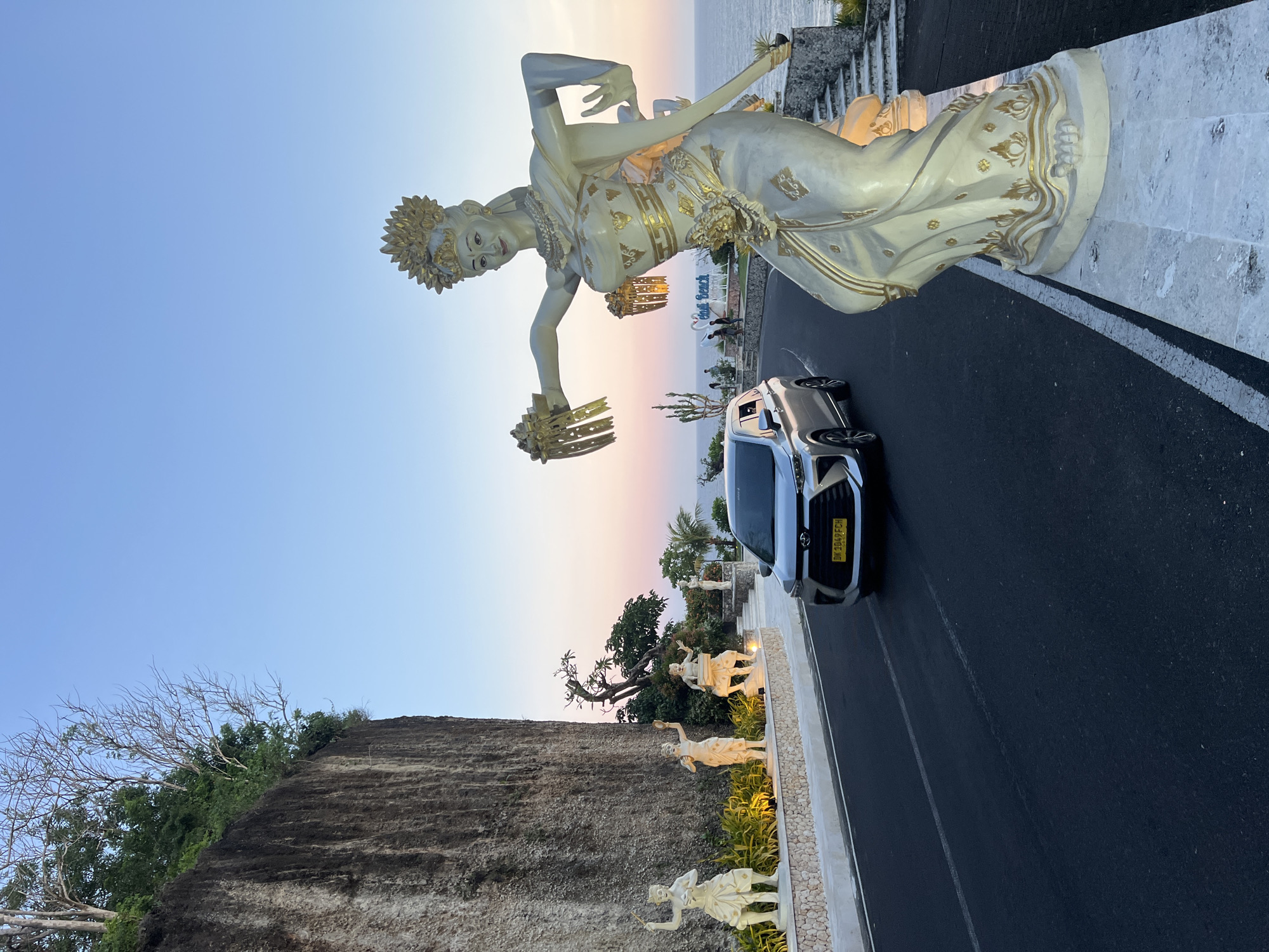 The image shows a road winding along a cliff side in Kuta Selatan, Indonesia. A large statue of a female dancer stands prominently at the roadside, overlooking the road and the ocean in the background. A silver car is parked on the road directly below the statue. Along the cliff side are smaller statues of people. The sky has a gradient of blue to pink, indicating either sunrise or sunset. The road is made of asphalt and has a white line marking the edge.
