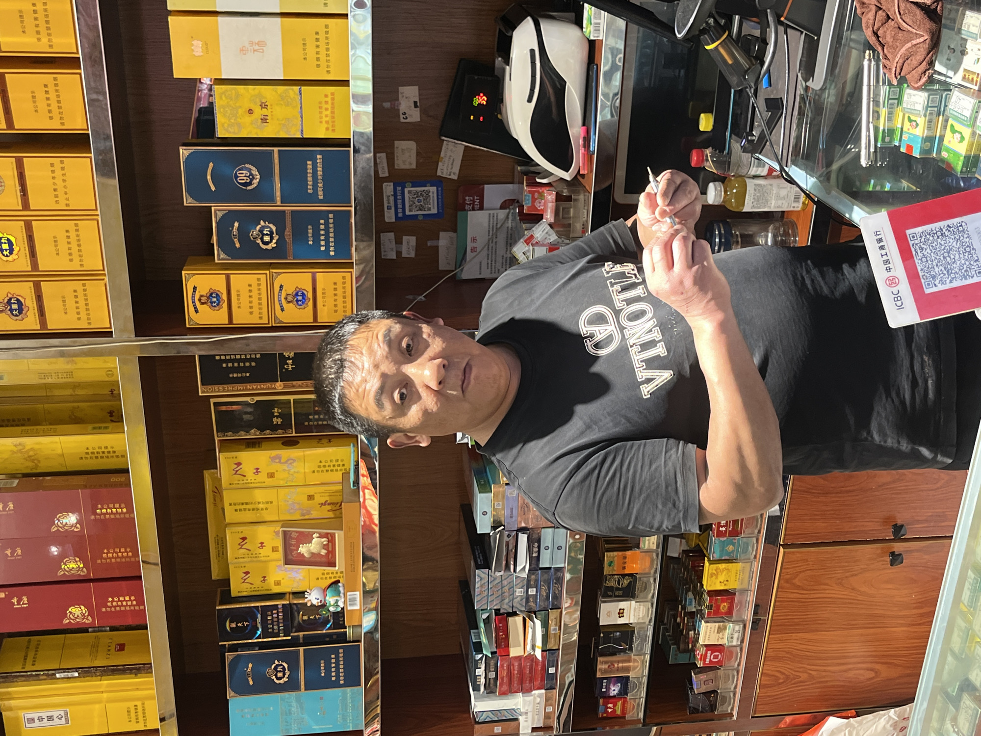 A person is holding a needle/syringe device, standing in front of shelves stocked with cigarettes. The shelves showcase multiple brands, including "Yunyan Impression", "Lion", "Tianzi", and "Chongyang". A red ICBC payment terminal is visible on the right. The location is suggested to be in Jiulongpo District, China, based on the Chinese characters and the ICBC terminal.