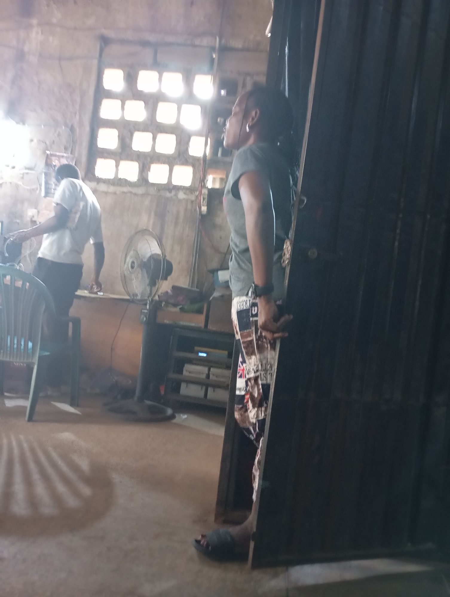 The image depicts an indoor scene, likely a workshop or storage area, in Shendam, Nigeria. A woman is standing in the foreground, facing left and looking towards the right. She has dark skin, dark hair pulled back, and wears a dark green t-shirt and patterned shorts. She has hoop earrings and a watch on her left wrist. Her right arm is bent at the elbow, and her hand is resting on her thigh. Behind her, a dark, tall, metal structure, possibly a large door or panel, occupies the right side of the frame.
In the middle ground, a man wearing a light-colored t-shirt and dark shorts is visible from the back, facing away from the camera. He appears to be working at a counter or table. To his left, a green plastic chair is partially visible. Further to the right, a black standing fan is positioned near a shelf unit that holds various items, including what looks like electronic devices. The background features a wall with a window made of square, translucent panes, allowing light to enter the space. The floor is concrete, and shadows are cast across it, suggesting artificial lighting and possibly the sun. The overall environment appears utilitarian and somewhat unkempt.