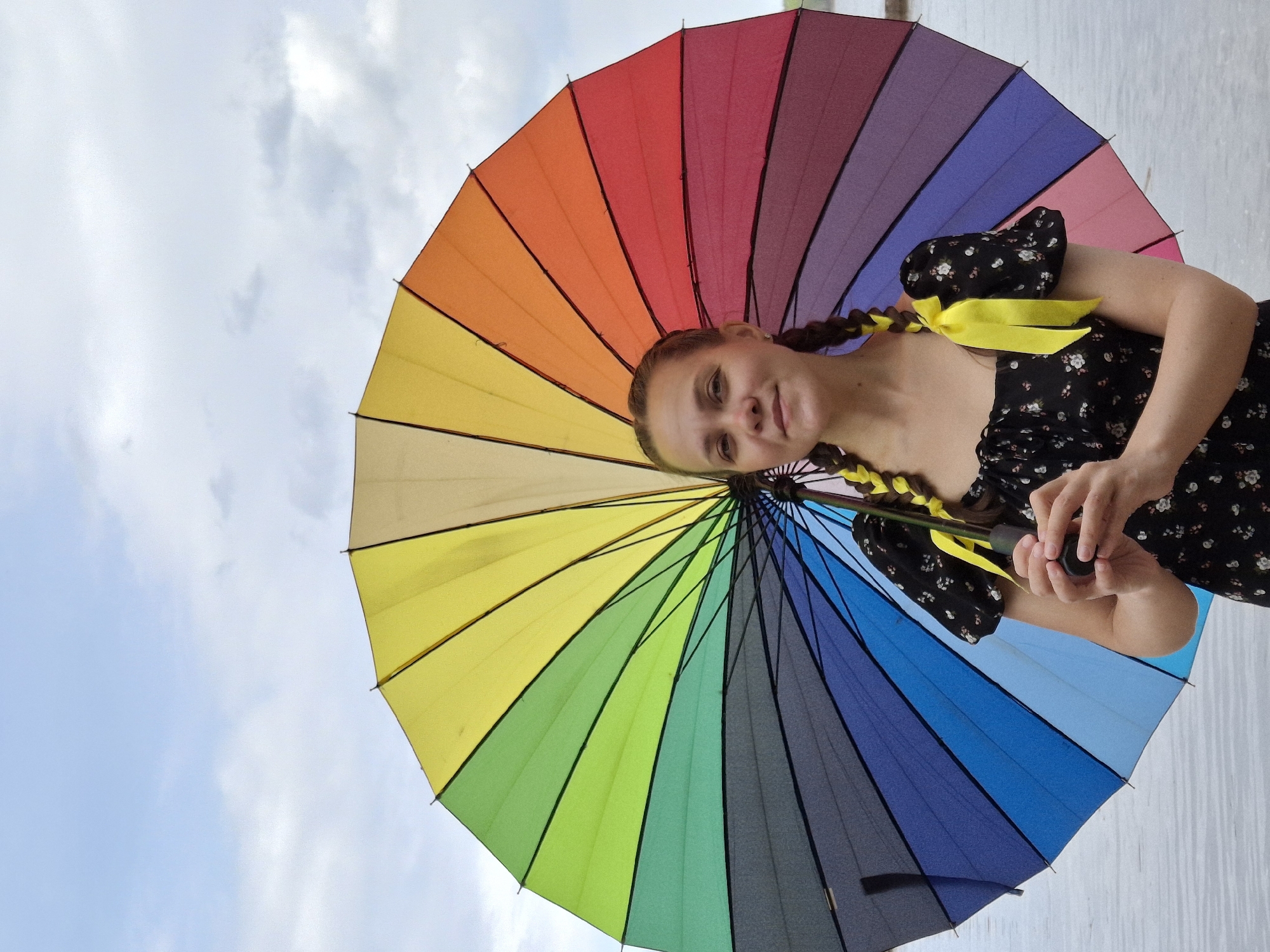 A fair-skinned young woman with dark hair braided and adorned with yellow ribbons poses under a large rainbow-colored umbrella. She is wearing a black floral-patterned dress with puffy sleeves. The umbrella's colors are arranged in a spectrum from red to purple, filling much of the frame. Parts of the sky, with scattered clouds, are visible behind the umbrella. The setting appears to be outdoors, with a white wall partially seen in the bottom right. The location is Unknown, Unknown.