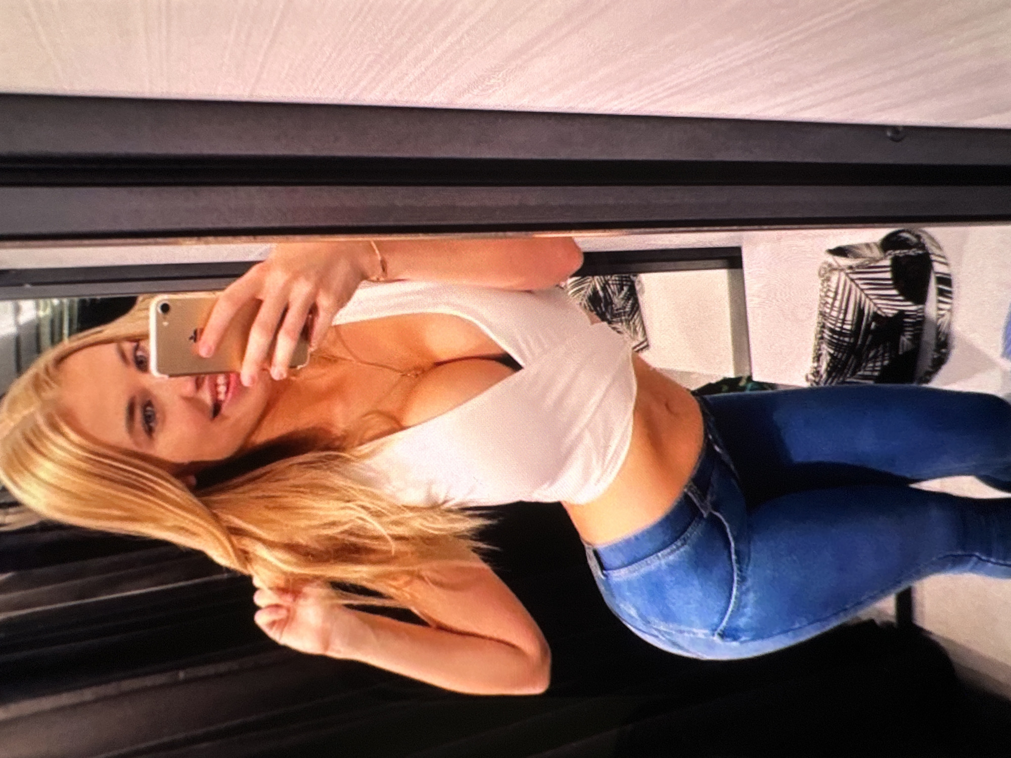 The media file shows a young woman with blonde hair taking a selfie in a mirror. She is wearing a white crop top and blue jeans. She is smiling and appears to be in a fitting room or a similar retail environment, as suggested by the presence of a mirror and dark, rippled curtains in the background. The lighting is bright, indicating daytime or well-lit indoor conditions. The woman is holding a gold smartphone in her right hand to capture the image. Her left hand is resting on her hair. The image is slightly tilted, adding a dynamic feel to the composition. The overall mood appears casual and confident.