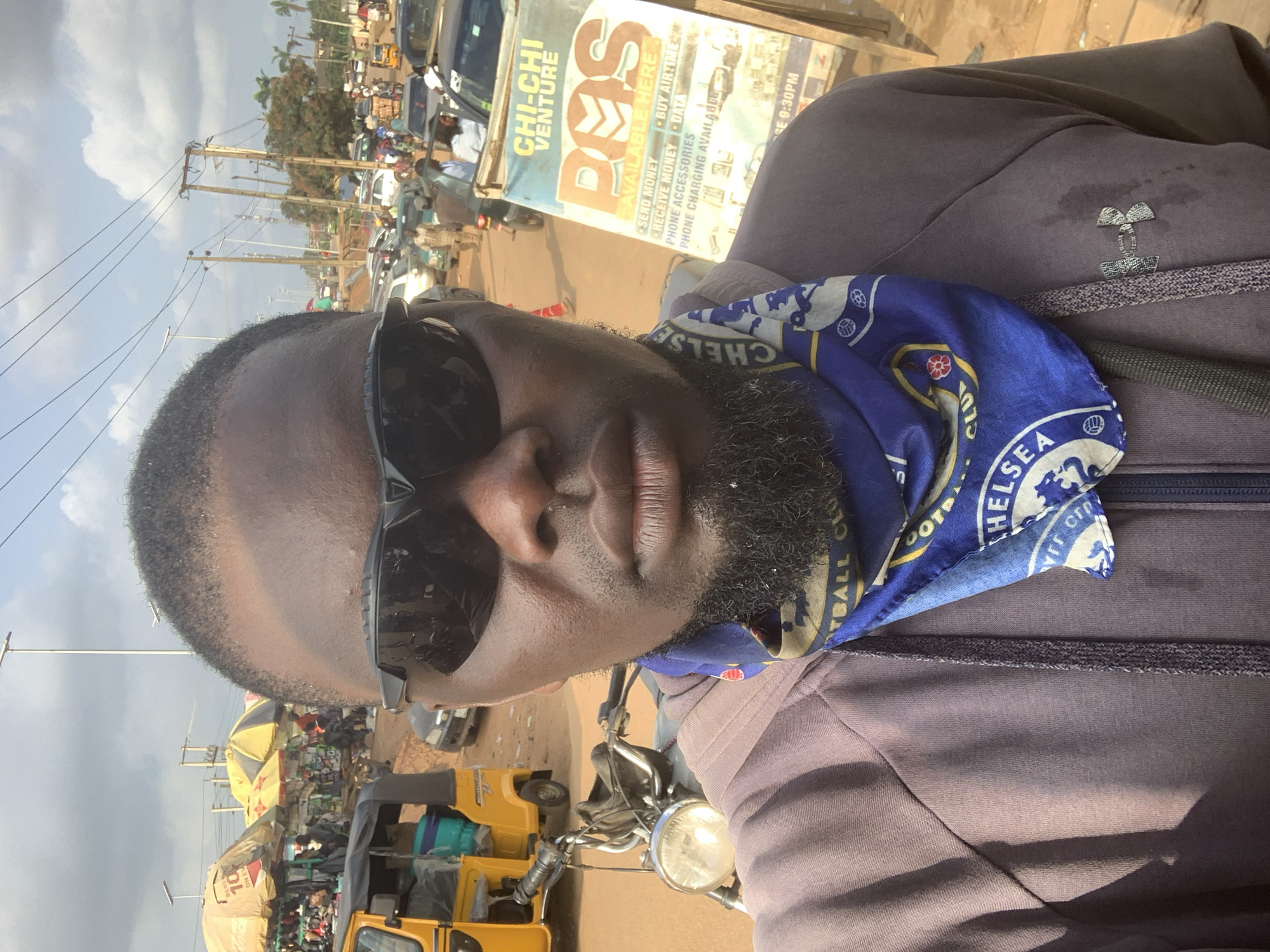 This is a selfie taken in Shagamu, Nigeria. The image features a dark-skinned man with sunglasses and a blue bandana around his neck. He is wearing a purple hoodie with an Under Armour logo on the right side. The background shows a busy street with several vehicles, including a yellow auto-rickshaw and cars. There are also market stalls with umbrellas and people milling around. A sign that reads "CHI-CHI VENTURE POS AVAILABLE HERE" is visible to the right of the man. The scene appears to be a commercial area with moderate traffic.