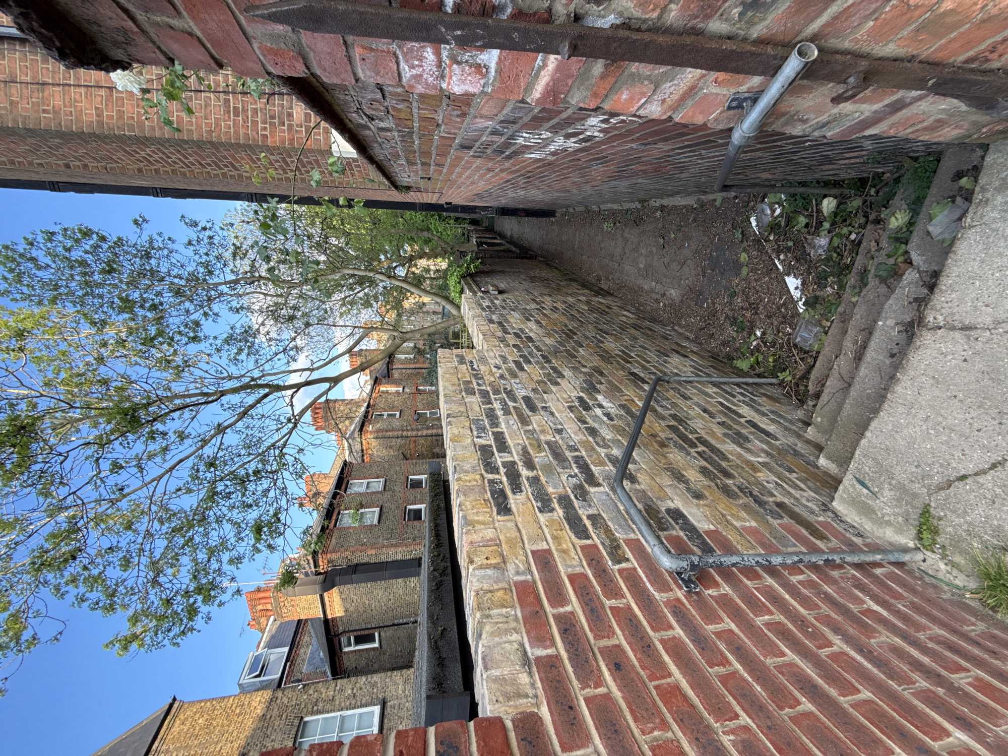 An outdoor alleyway in an urban environment, potentially London, United Kingdom. Brick walls define the narrow space on either side, with a concrete path running in the middle. A pipe runs along the brick wall to the right, and another pipe is installed as a handrail along the left side of the path. Overhanging the walls is a tree with bare branches. Several buildings are located behind the brick wall on the left. Litter and sparse vegetation are seen on the path. The sky is visible at the top-left of the frame.