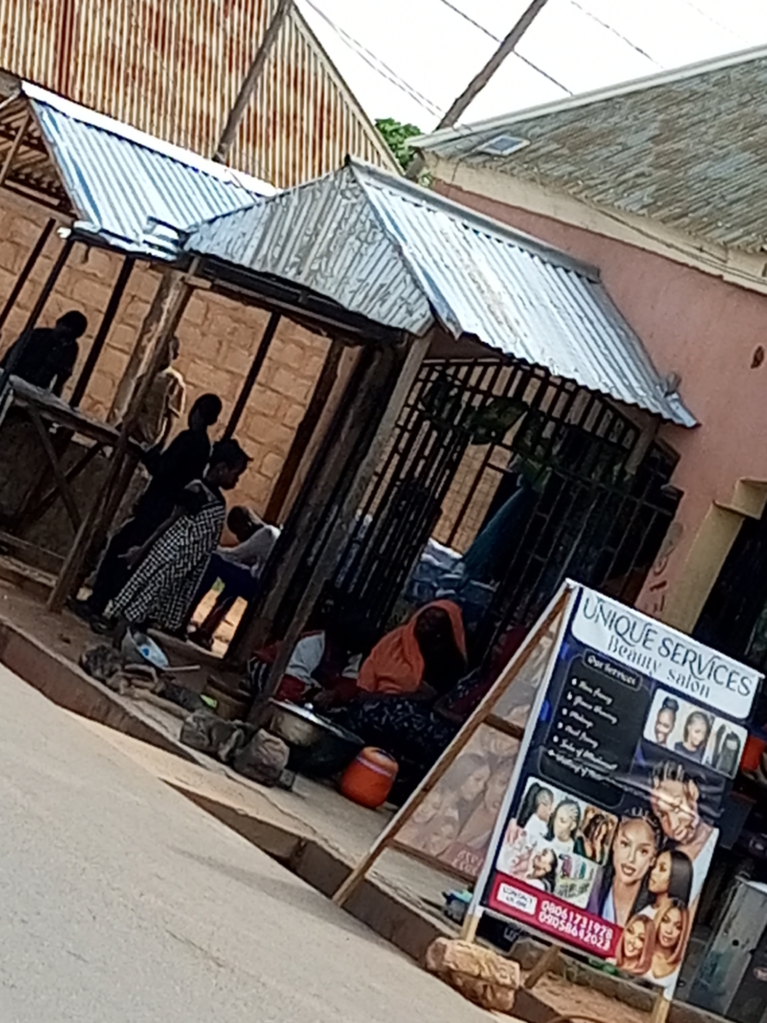 A low-angle, eye-level, medium shot captures a street scene in Tempure, Nigeria, characterized by a mix of commercial and residential structures. The foreground shows a light-colored, slightly textured pavement bordering a raised, reddish-brown walkway. To the left of the walkway, an orange bucket sits near some grey, irregular-shaped objects, possibly stones or small rocks.
Dominating the mid-ground is an open-fronted structure with a corrugated metal roof supported by wooden posts. This structure appears to be a shop or stall, with a brick wall behind it. Several individuals are visible within and around this structure. In the foreground of the stall, a person in an orange headscarf and dark clothing is seated, facing away from the camera. To their left, another individual is partially visible, wearing a white and black checkered garment. Further back, silhouetted figures are seen standing and sitting.
In the right mid-ground, a standing signpost displays "UNIQUE SERVICES Beauty salon" at the top. Below this heading, a list of services includes "Our Services," "Hair perming," "Ghana Weaving," "Makeup," "Facial," and "Nail of Mankind." The sign also features images of various hairstyles and faces of women, and contact information including phone numbers "08061731978" and "09058642023." The signpost is angled towards the viewer and is supported by a wooden easel.
The background reveals more of the environment, including buildings with corrugated metal roofs, one with a distinctive rusty, ribbed texture, and another with a lighter, more weathered corrugated surface. The overall impression is one of a busy, informal commercial area, likely a street market or roadside business district, common in Nigerian cities. The lighting suggests it is daytime, with diffuse light.