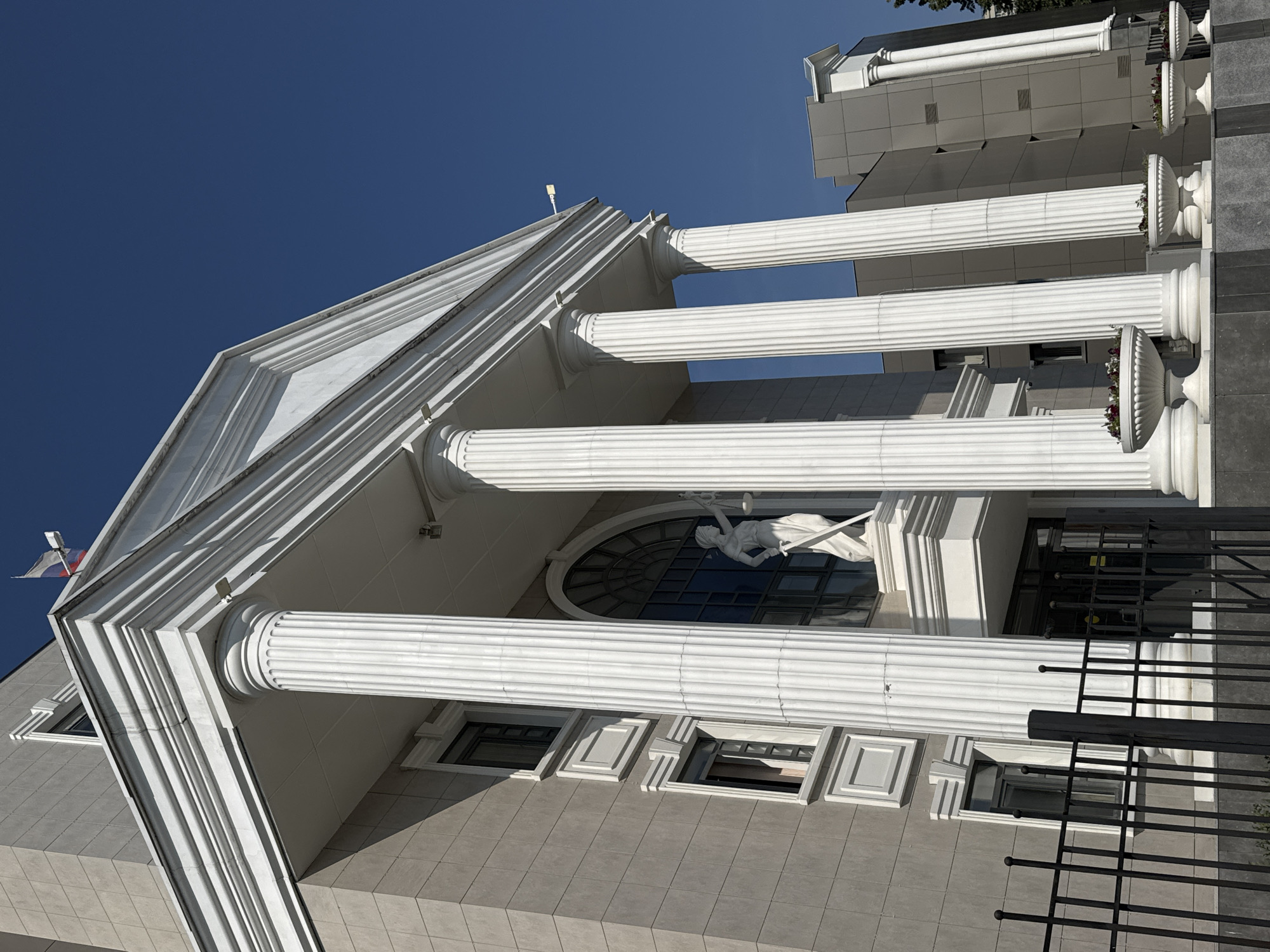 An exterior, eye-level, medium shot captures a building with a neoclassical facade against a clear, deep blue sky. The building is predominantly grey, with white columns and architectural embellishments. A statue of Lady Justice, holding a scale and sword, stands at the entrance. The statue is white and positioned on a pedestal above a set of doors. Multiple large, fluted white columns support a triangular pediment. The facade also features several windows, some rectangular and others part of a large arched window behind the statue. In front of the building, a black metal fence runs horizontally across the lower portion of the frame. Several white planter urns with foliage are situated along the base of the building. In the background, a modern grey building is visible, partially obscured by the neoclassical structure. A Russian flag flies from a pole on the roof of the neoclassical building. A street light is visible on the upper right of the neoclassical structure.