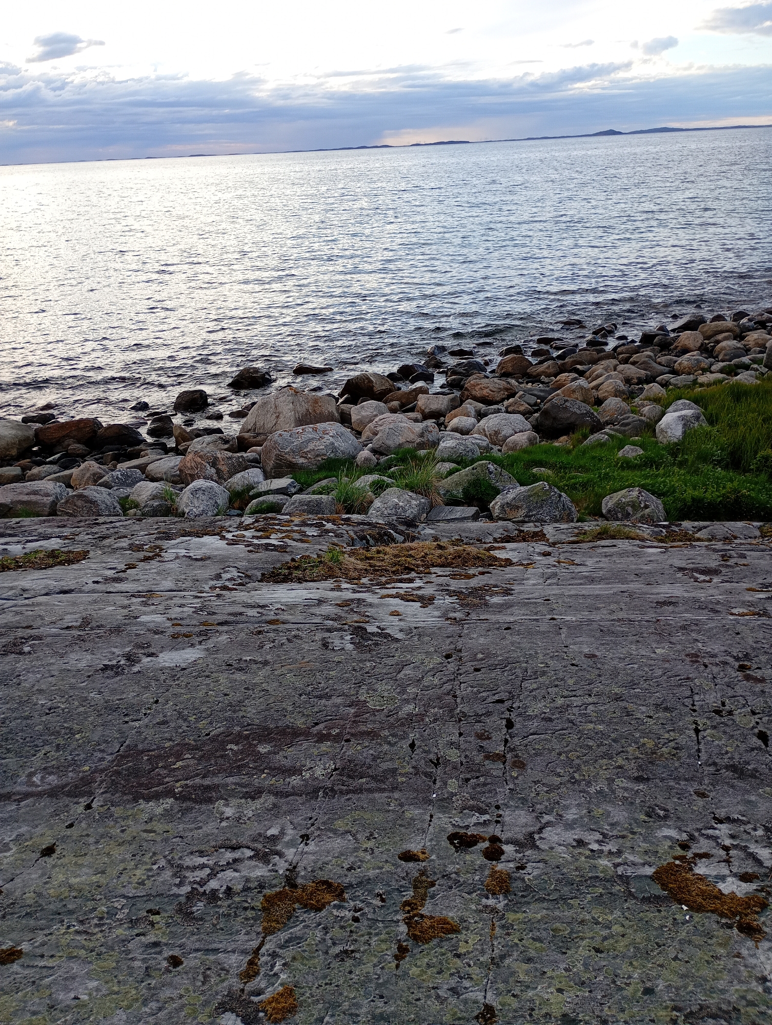 A natural scene shows a rocky shoreline meeting a calm body of water under a cloudy sky. The foreground is dominated by a large, textured gray rock with patches of moss and dry grass. Behind this rock, the shore is covered with numerous rounded stones of various sizes, ranging from small pebbles to larger boulders. Some patches of green grass and vegetation are visible between the stones. The water is dark and rippling, reflecting the light from the overcast sky. In the distance, the horizon is marked by a faint outline of land. The overall impression is one of a rugged, natural coastal environment.