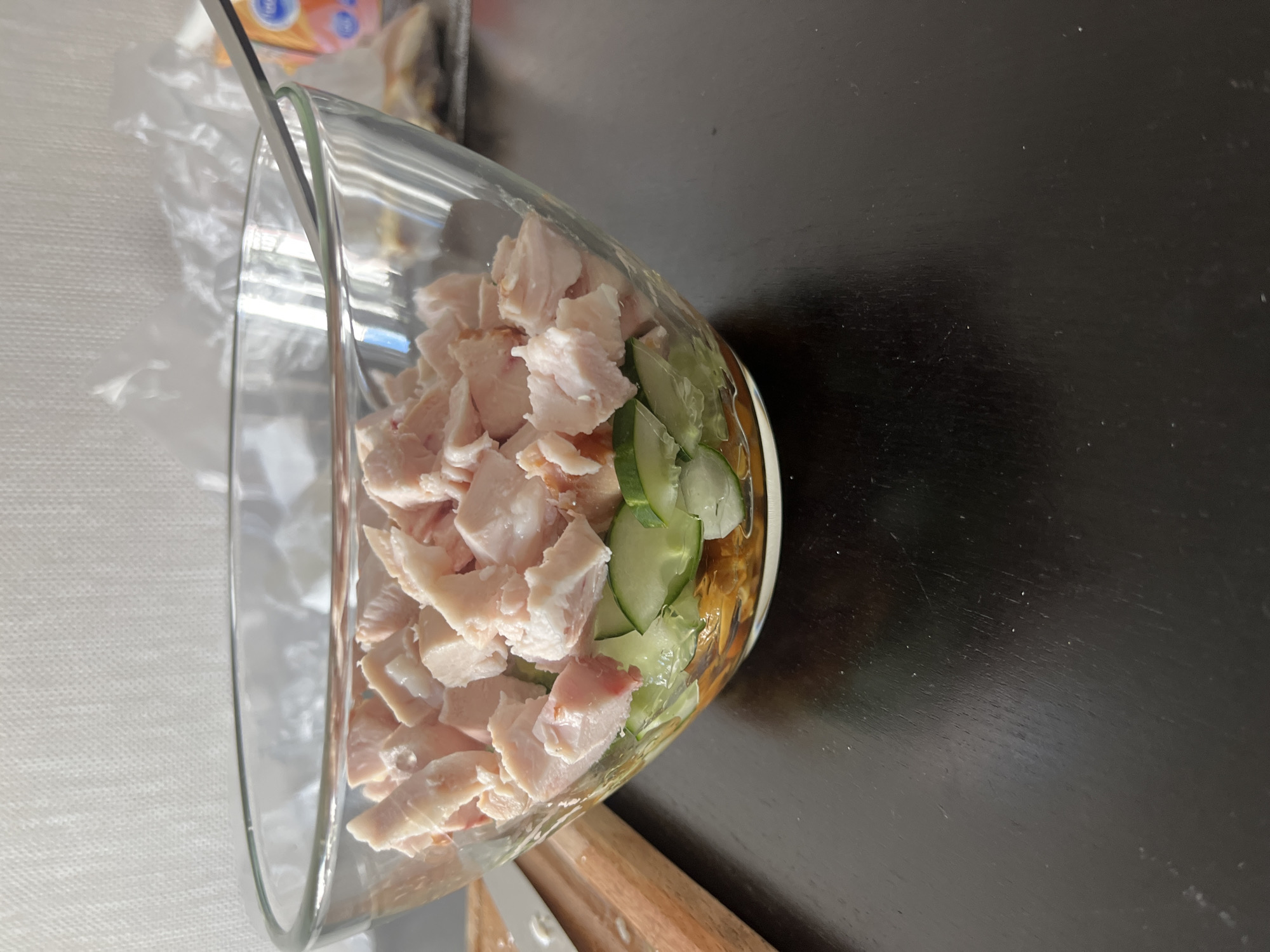 A close-up, eye-level shot captures a transparent glass bowl filled with a layered salad. The salad's top layer consists of bite-sized pieces of cooked meat, likely chicken or pork, with some visible fat. Beneath the meat, sliced cucumber rounds are arranged, and further down, a layer of what appears to be finely shredded carrots or another orange vegetable is visible. A fork is resting on the rim of the bowl. The bowl sits on a dark, reflective surface, possibly a wooden table, with a wooden cutting board and a knife visible to the left of the bowl. In the background, a white textured wall is present, along with some crumpled plastic packaging. The overall lighting suggests an indoor setting, likely a kitchen or dining area. No specific location in Perm, Russia can be identified from the image.
