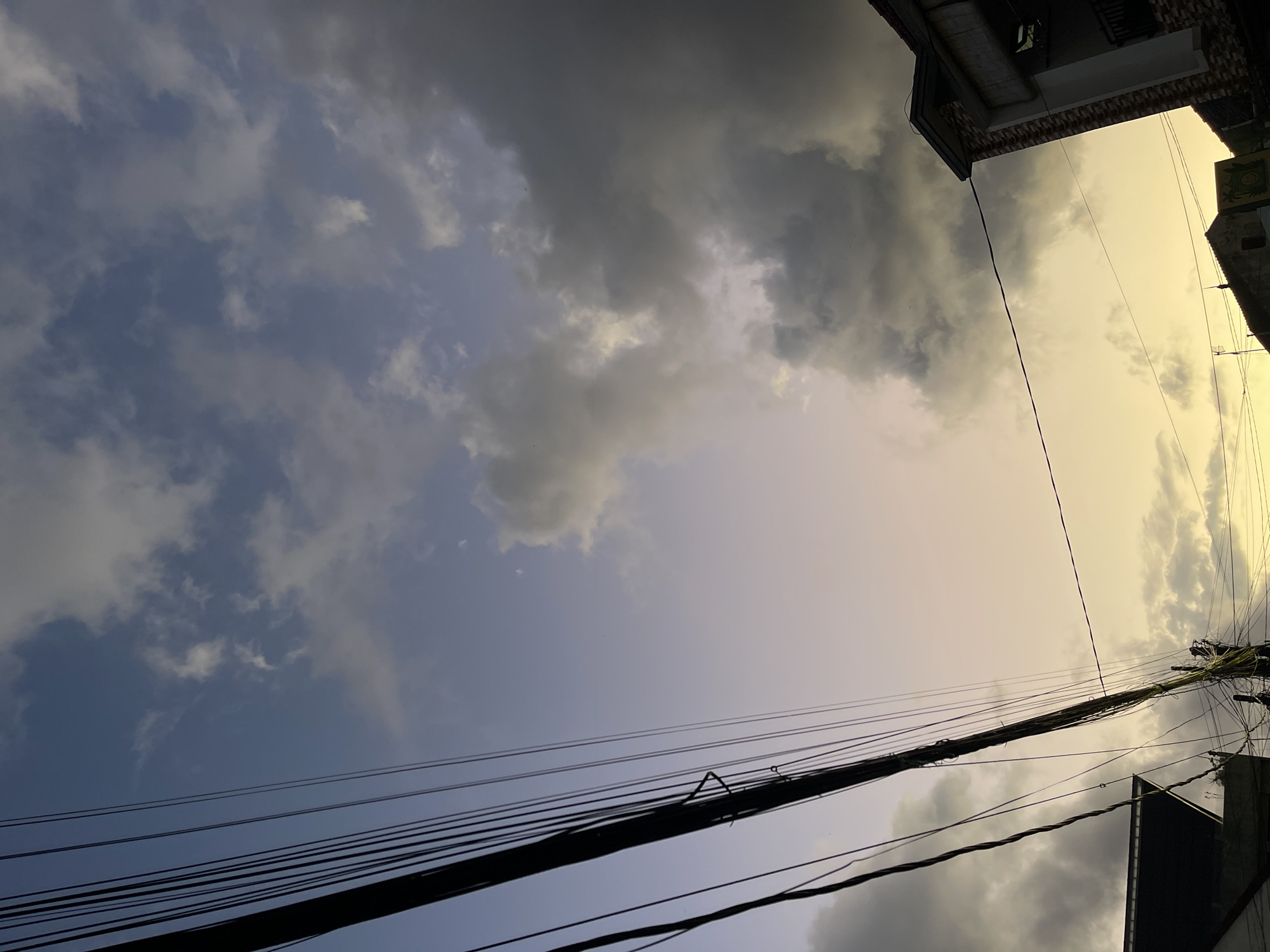The image captures a view of the sky above North Kuta, Indonesia, at dusk. A dense network of black electrical wires dominates the left side of the frame, stretching diagonally across the scene. These wires are attached to a wooden pole visible in the lower right quadrant. To the left of the wires, the dark roofline of a building is partially visible. On the right side of the image, the corner of a modern building with brick and white trim is present, featuring a balcony. The sky is a dramatic mix of colors, with the upper portion showcasing a clear blue with scattered white clouds, transitioning to a soft yellow and pale purple gradient towards the horizon, where darker, storm-like clouds gather on the right. The overall impression is of a city landscape under a changing sky.