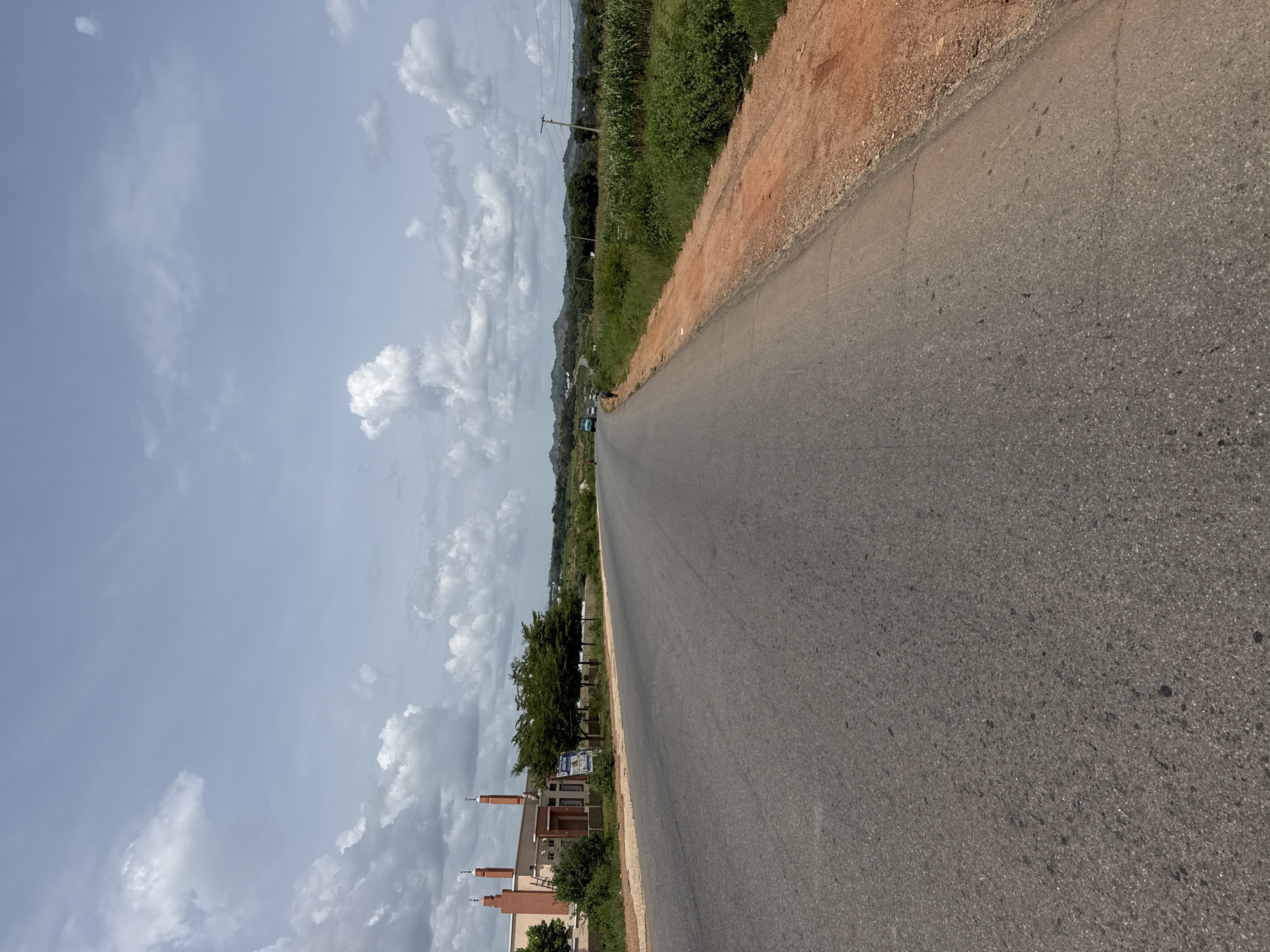 An asphalt road stretches into the distance, curving gently to the right. On the left side of the road, a light-colored building with red-tiled roofs and minarets is visible, suggesting it might be a mosque. Several trees stand between the building and the road. The road is bordered on the right by a reddish-brown dirt shoulder and then a field of green vegetation, including what appears to be a cornfield. In the background, rolling hills are dotted with trees and scattered structures. The sky is partly cloudy with fluffy white clouds against a pale blue backdrop. A few vehicles and pedestrians can be seen on the road in the distance, indicating some activity.