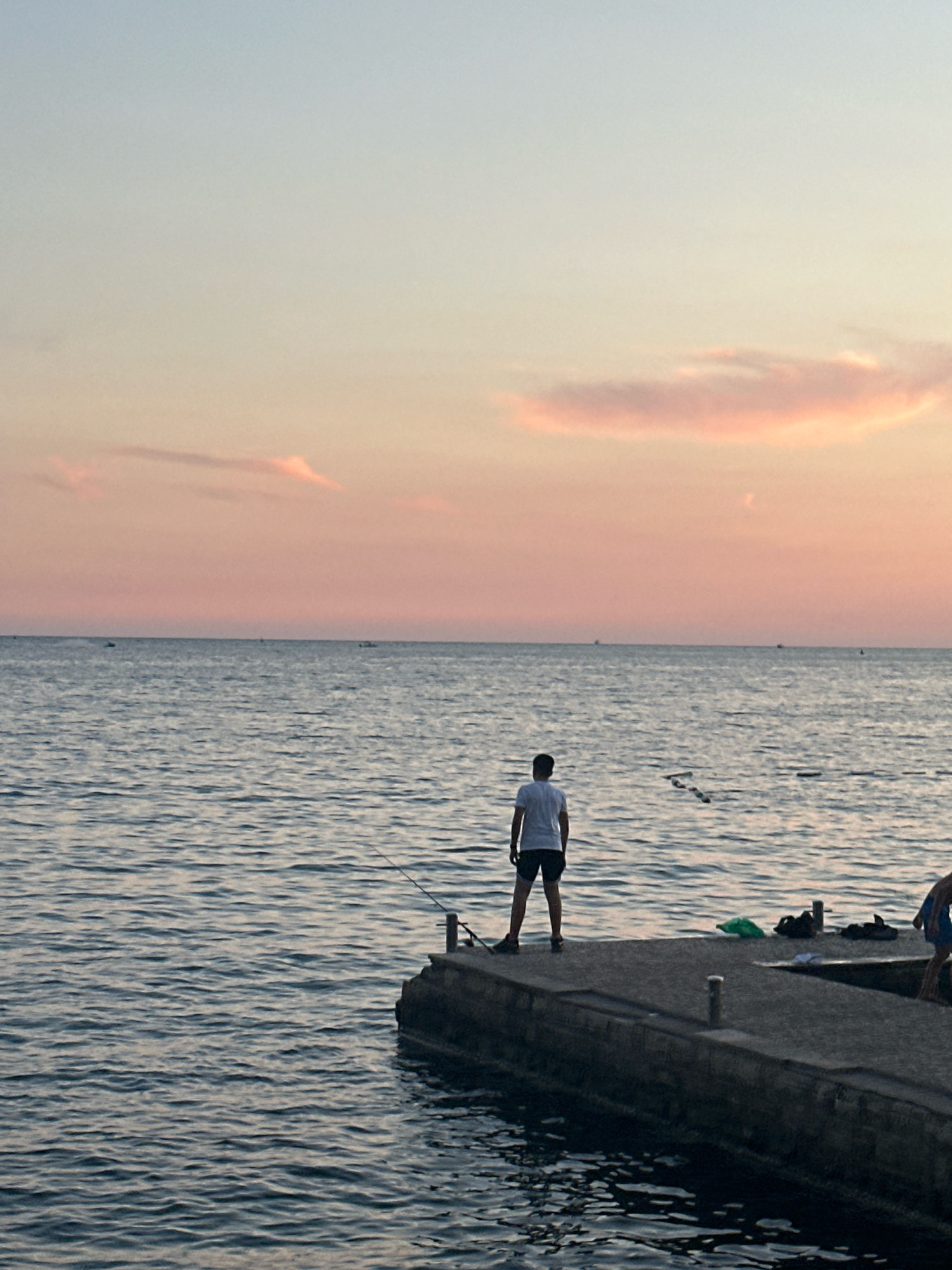 A lone fisherman stands on a concrete pier at dusk, facing the calm sea. The sky above is a soft gradient of pale blue and pink, with wisps of clouds catching the last rays of sunlight. The water is a deep blue, rippling gently. In the distance, several small boats are visible on the horizon. The pier extends into the water, and on it, near the fisherman, are a green bag and a pair of sandals. Another person, only partially visible in the lower right corner, is also on the pier, facing away from the camera and towards the water. The overall scene suggests a peaceful evening by the sea. The location is likely the coast of Budva, Montenegro, given the typical coastal landscape and the style of the pier.
