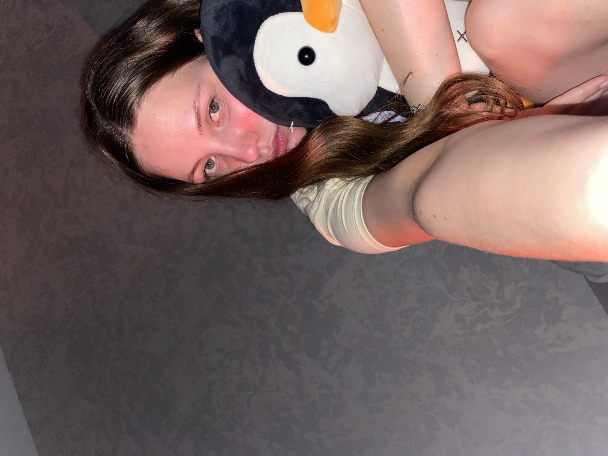 A young woman with long brown hair is holding a stuffed penguin in her arms. She is looking at the camera with a neutral expression. Her left cheek is flushed red, possibly from lighting or skin irritation. The background is a dark, textured wall. The lighting is dim and appears to be coming from a red light source. The scene is likely indoors, in a residential setting. There is no information in the image to suggest a specific location in Izhevsk, Russia, or any other city.