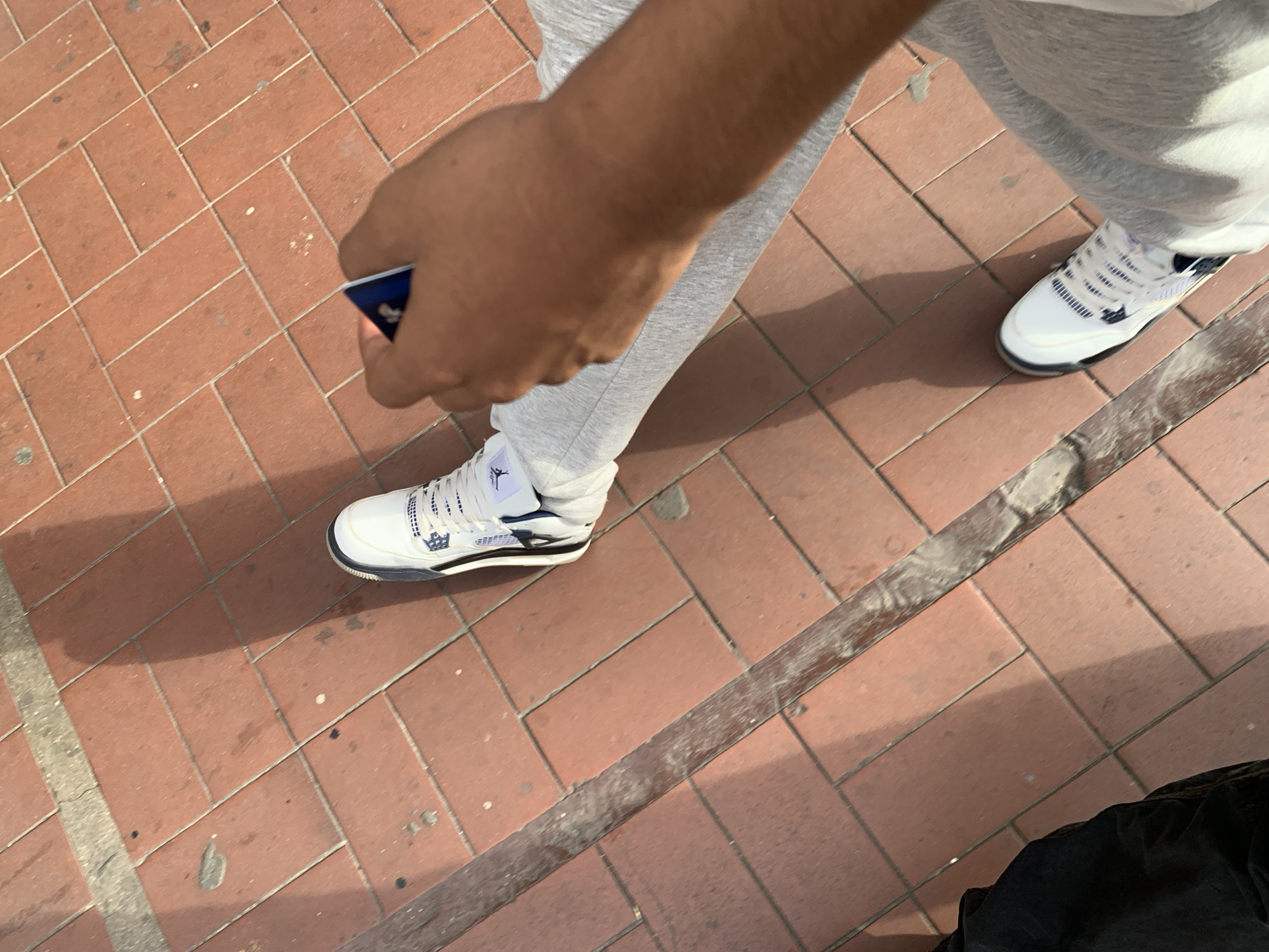 The image depicts a ground-level view of two people walking or standing on a terracotta-colored brick pavement in an outdoor setting, likely an urban area in Porlamar, Venezuela, during daylight hours. The pavement consists of rectangular bricks laid in a patterned arrangement.
In the foreground, a person's right arm and hand are visible, holding a small blue card. This individual wears grey sweatpants and a white, blue, and black Air Jordan 4 sneaker, featuring the iconic Jumpman logo prominently on the tongue. Strong, distinct shadows are cast by the person's legs and arm onto the pavement, indicating bright, direct sunlight and clear weather.
Further back and to the right, the leg and foot of a second person are visible, dressed in similar grey sweatpants and a matching white, blue, and black Air Jordan 4 sneaker. A long, weathered metallic bar or railing stretches diagonally across the lower half of the frame, partially obscured by shadows. The scene suggests a casual moment outdoors on a sunny day. The only visible text is the Jumpman logo on the sneakers.