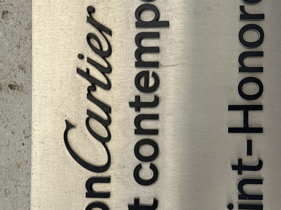 The image displays a close-up, frontal view of a rectangular, horizontally brushed metal plaque, likely made of stainless steel or aluminum, affixed to a lighter-colored, textured stone or concrete wall. The plaque features three lines of raised, black, three-dimensional lettering. The top visible line reads "...on Cartier," with "Cartier" rendered in a distinctive script font. The middle line displays "t contemp," in a sans-serif block font, consistent with "art contemporain" (contemporary art). The bottom line shows "int-Honor," also in a sans-serif block font, consistent with "Saint-Honoré." The plaque shows minor surface wear and vertical streaks consistent with water residue or grime. The combination of "Cartier," "contemporary art," and "Saint-Honoré" indicates this sign belongs to a Cartier establishment, such as a boutique or exhibition space, located on or in the vicinity of Rue Saint-Honoré in Paris, France. No individuals, actions, or interactions are present in the scene.