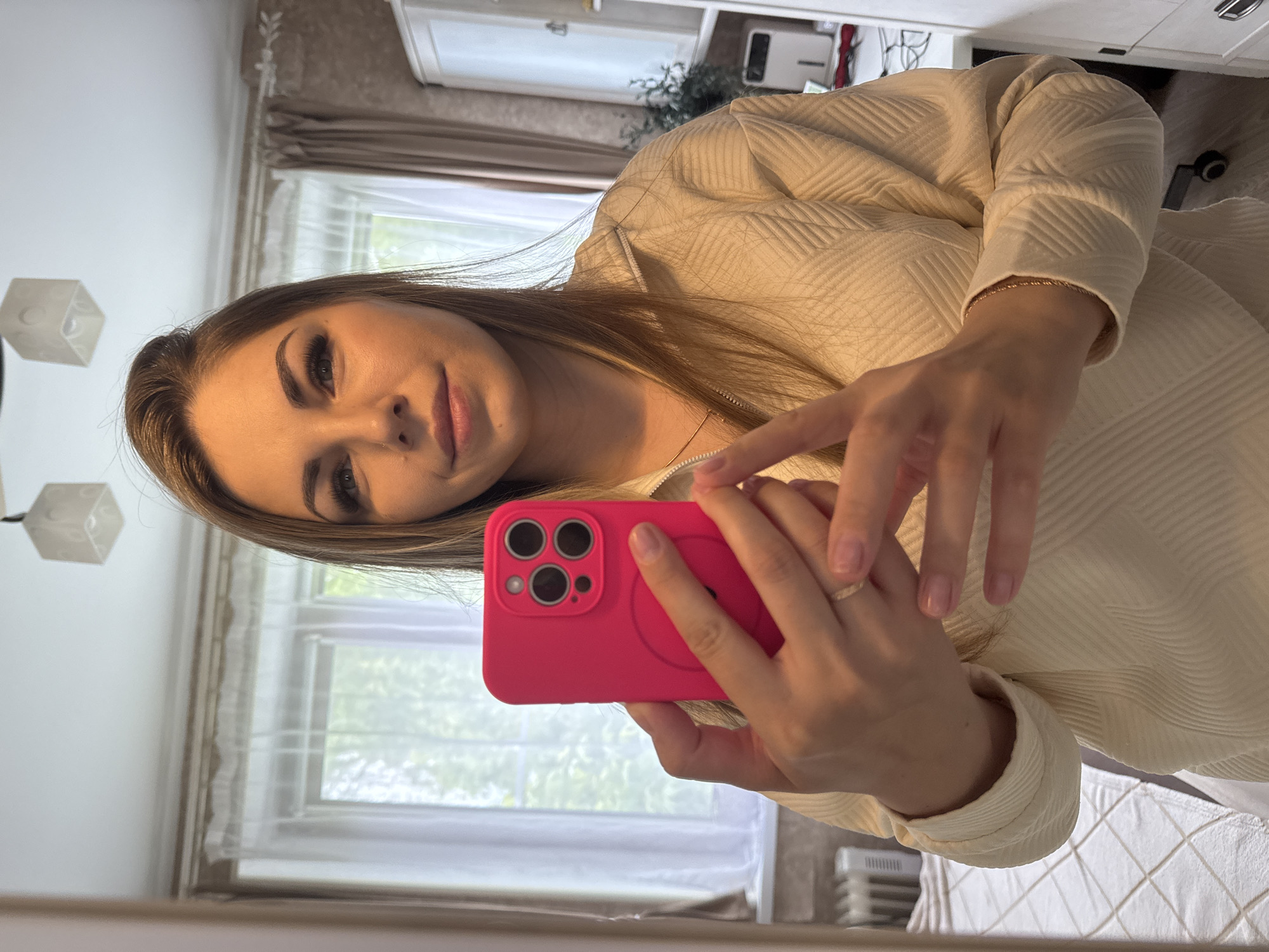 A woman is taking a selfie with a bright pink smartphone. She is indoors, possibly in an apartment. The background shows a window with sheer curtains and a view of greenery. The woman is wearing a light-colored, textured sweater. A lamp is visible on the left side of the image, and a cabinet is visible behind the woman. The location appears to be in Razdolye, Russia based on the prompt.